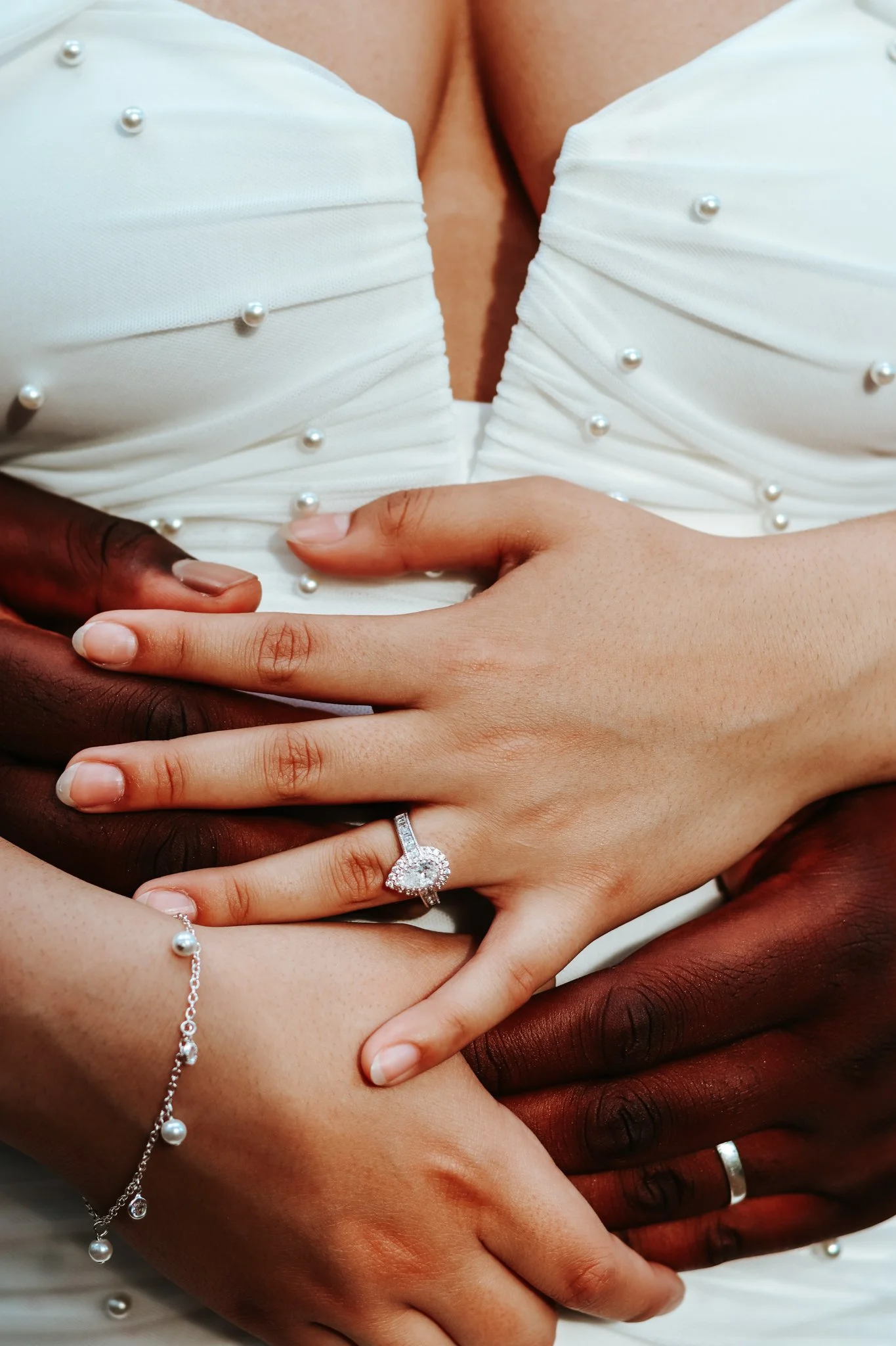 Close-up of intertwined hands of diverse individuals with wedding and jewelry, symbolizing unity and love, set against a background of wedding attire.