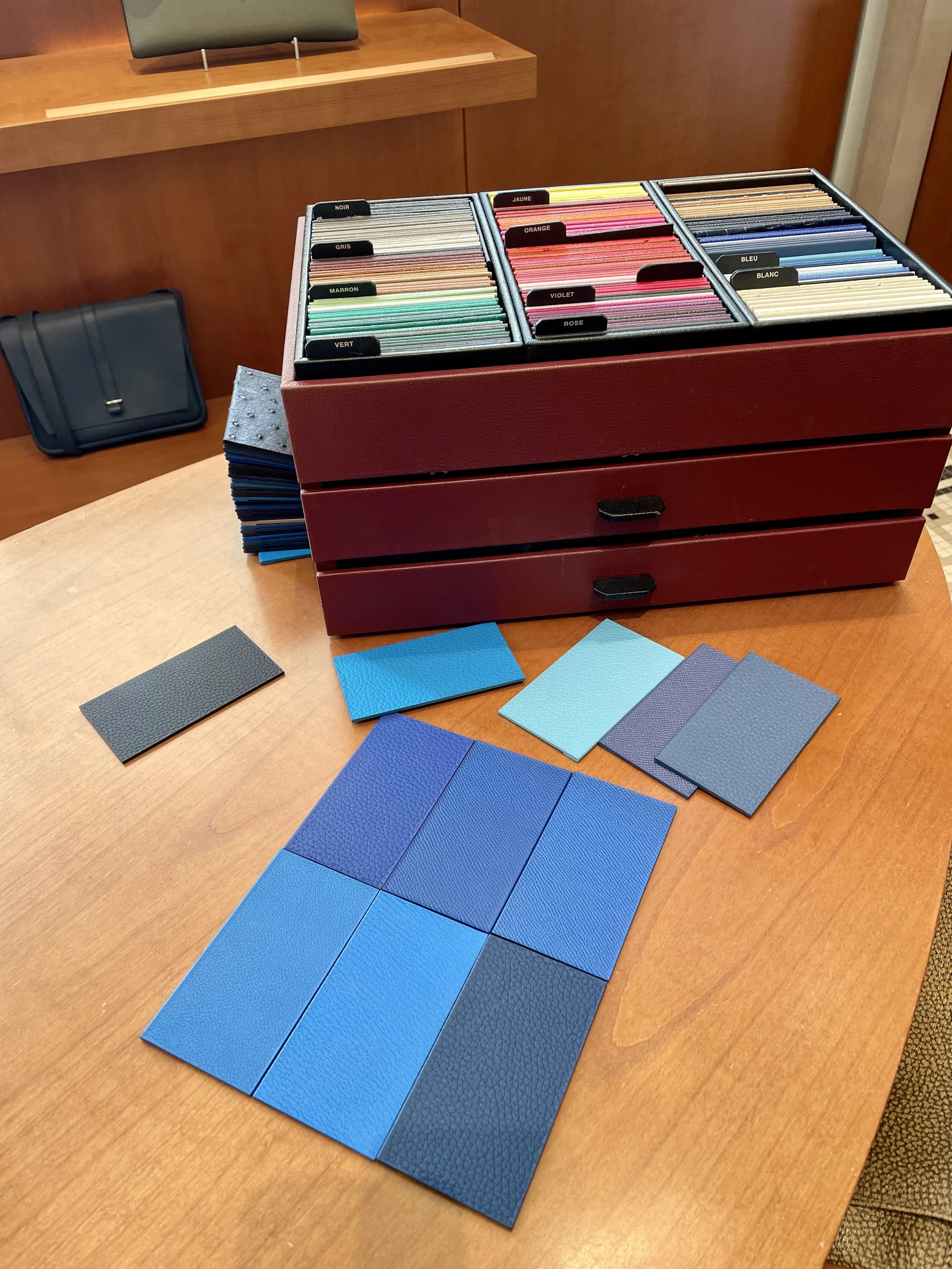 Samples of textured and solid-colored materials for a Hermes Birkin arranged on a wooden table in shades of black, blue, lFabric and leather swatches laid out on a desk during a sourcing session with Dubai personal stylist Manuela Cavallero