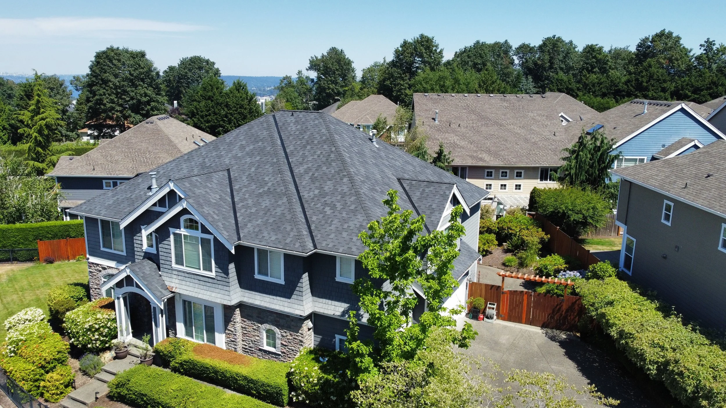Aerial view of a suburban house with a dark gray roof and landscaped garden surrounded by other homes and trees.