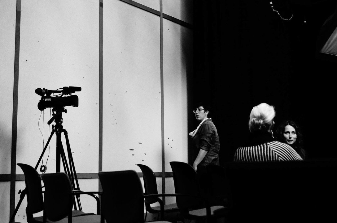 Sarah Klein, a white woman with short brown hair and glasses, walks toward a camera on a tripod. There are black stacking chairs visible in the foreground. Julie Burelle is visible speaking with an audience member with light hair and a striped sweate