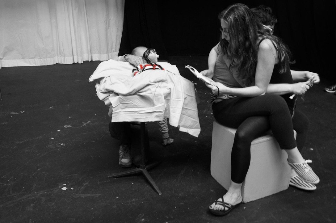 Black and white photo of Sarah Klein and Yelena Gluzman sitting on a white stage cube, legs scissored together on one another's laps. . Sarah knitting behind Yelena's back. Yelena is holding an open book behind Sarah's back. Yecid Calderon Rodeo is c