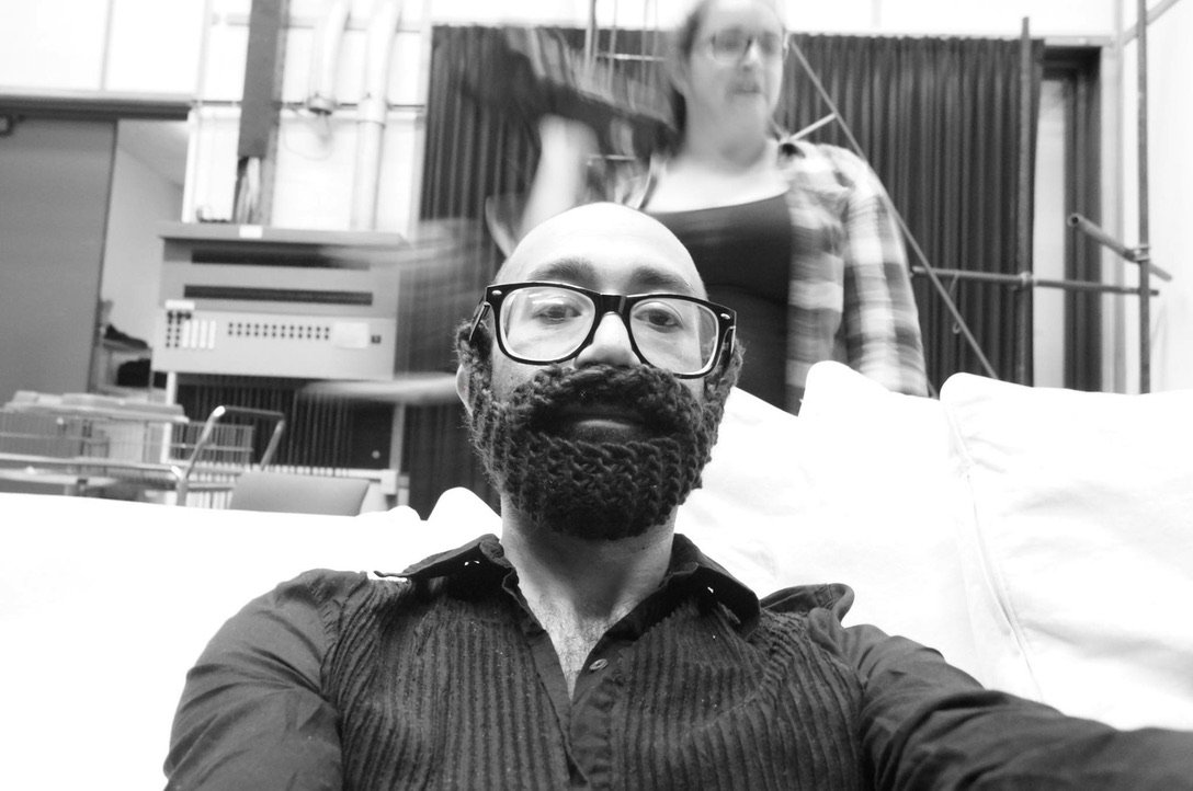 Yecid Calderon, visible from the chest up, is sitting on a light coloured sofa. He wearing a dark button down shirt and a knitted beard over his own beard. Christina Aushana gestures blurrily in the background.