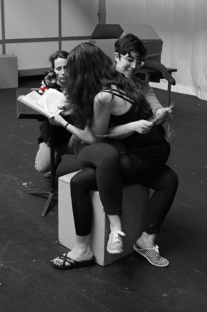 Black and white photo of Sarah Klein and Yelena Gluzman sitting on a white stage cube, legs scissored together on one another's laps. Sarah is knitting behind Yelena's back and Yelena is reading from a book. Julie Burelle is partially visible crouchi