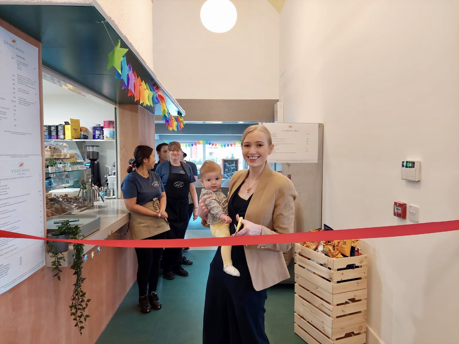 Robyn about to cut a red ribbon to mark the opening of the Shinfield Robyn Nest in 2021. She is smiling and holding her 6 month old daughter with some female staff members smiling behind her.