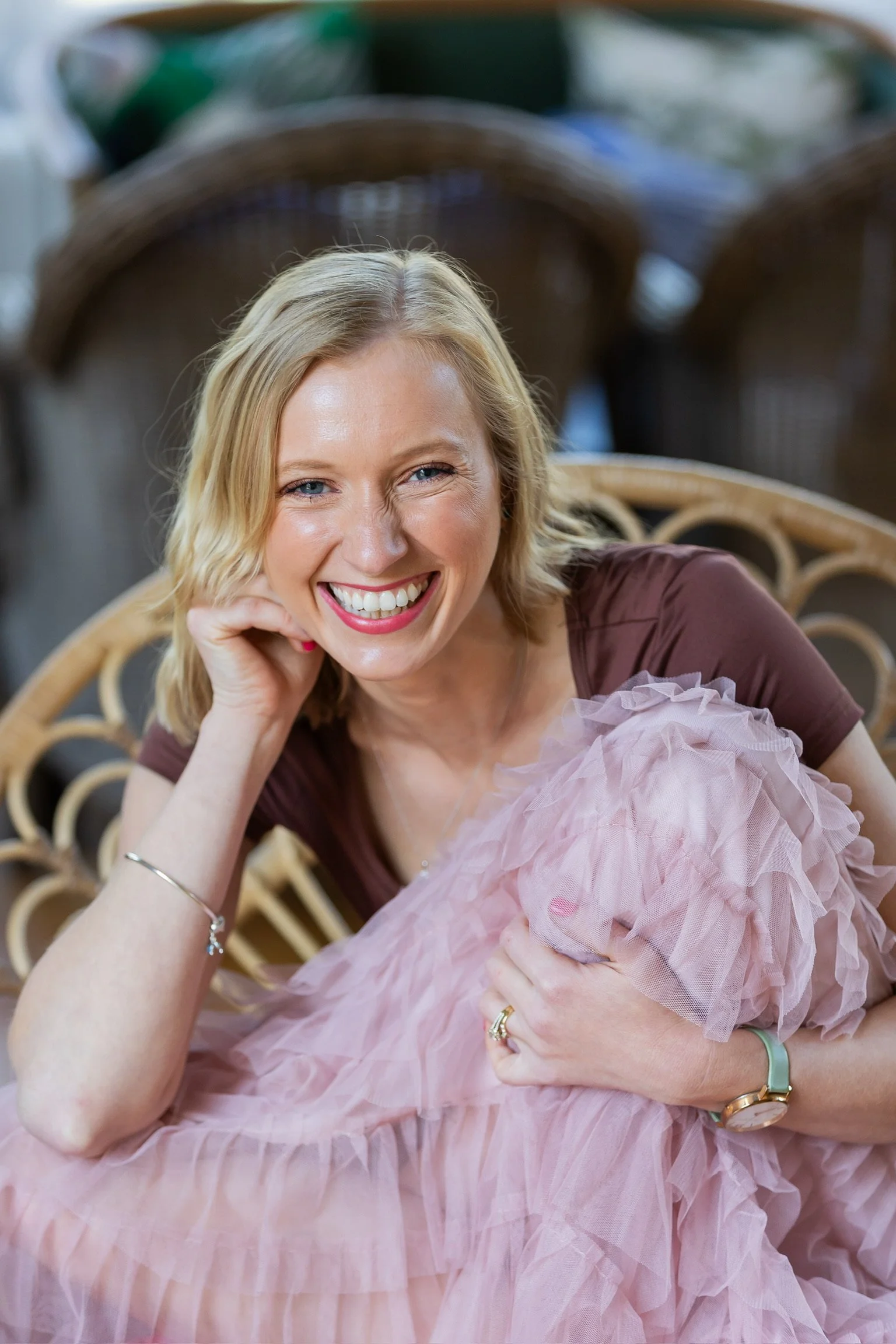 Robyn with a wide genuine warm smile sat in a peacock chair hugging her knee in a pink tutu style skirt and brown tshirt.