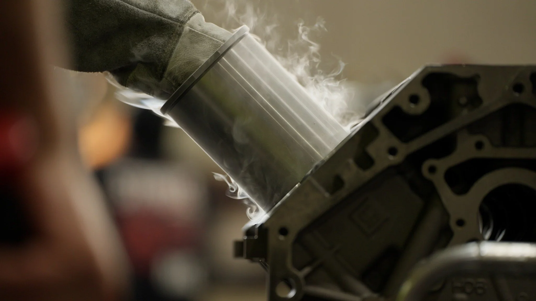 A close-up of a person handling a metal cylinder emitting smoke or vapor, positioned next to an engine block with visible cylinder head or engine parts.