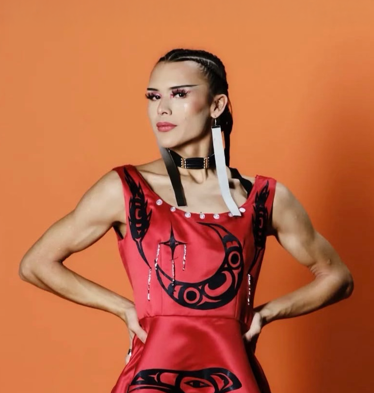 A woman in a patterned black and red dress standing against an orange background at Vancouver Indigenous Fashion Week.