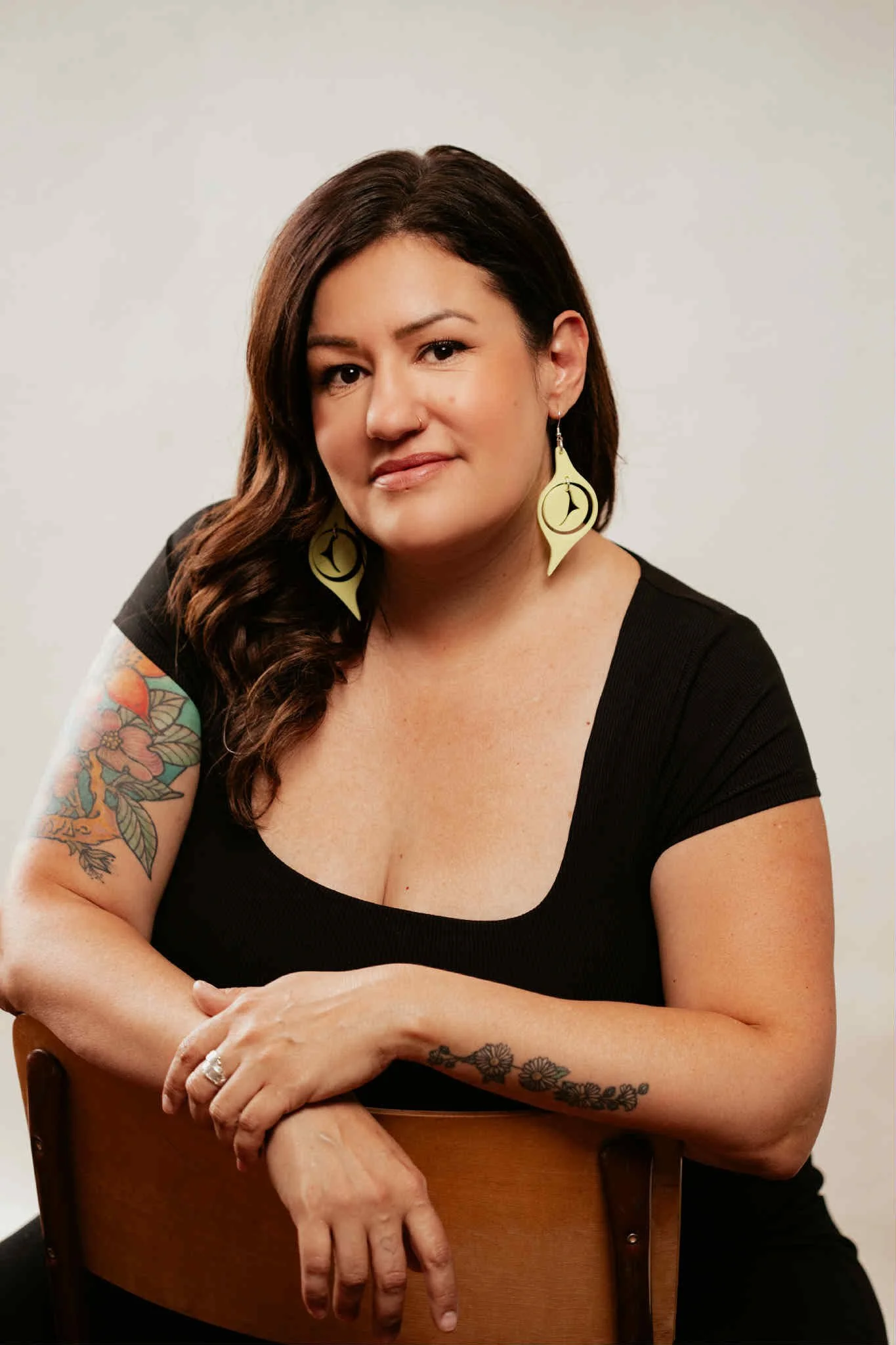 A woman with shoulder-length, wavy dark hair, wearing large, cage-style earrings, a black top, and a black leather jacket, smiling at the camera against a plain white background.