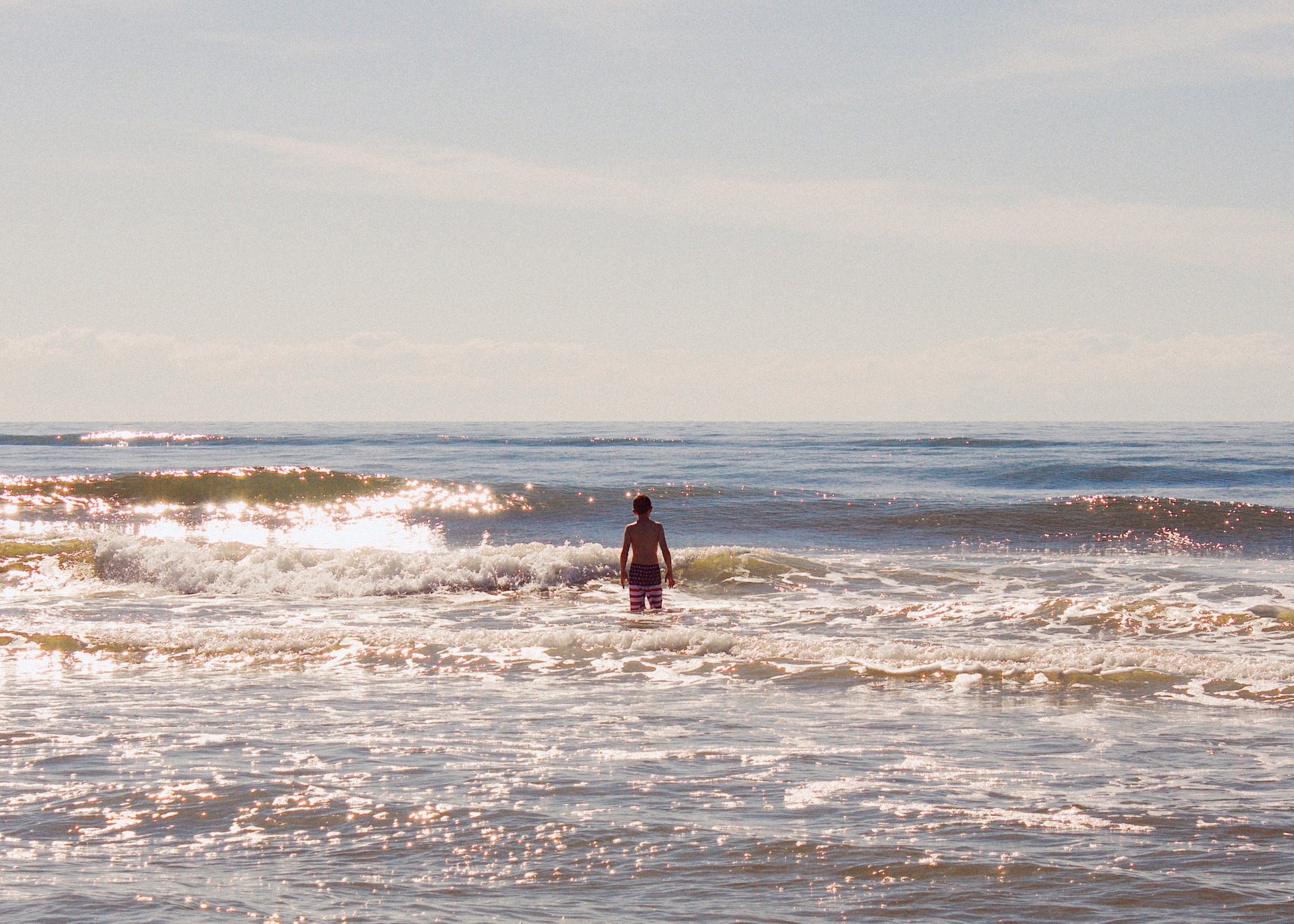 A boy standing in the ocean waves during daytime, seen from behind, with a clear sky and horizon in the background.