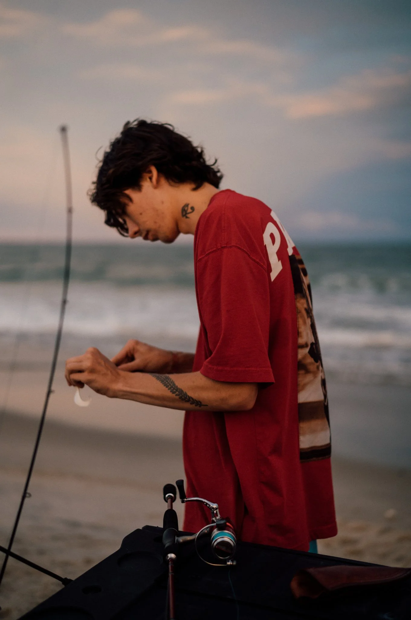 A young man with tattoos on his arm and neck, wearing a red T-shirt, preparing fishing gear on the beach at sunset.