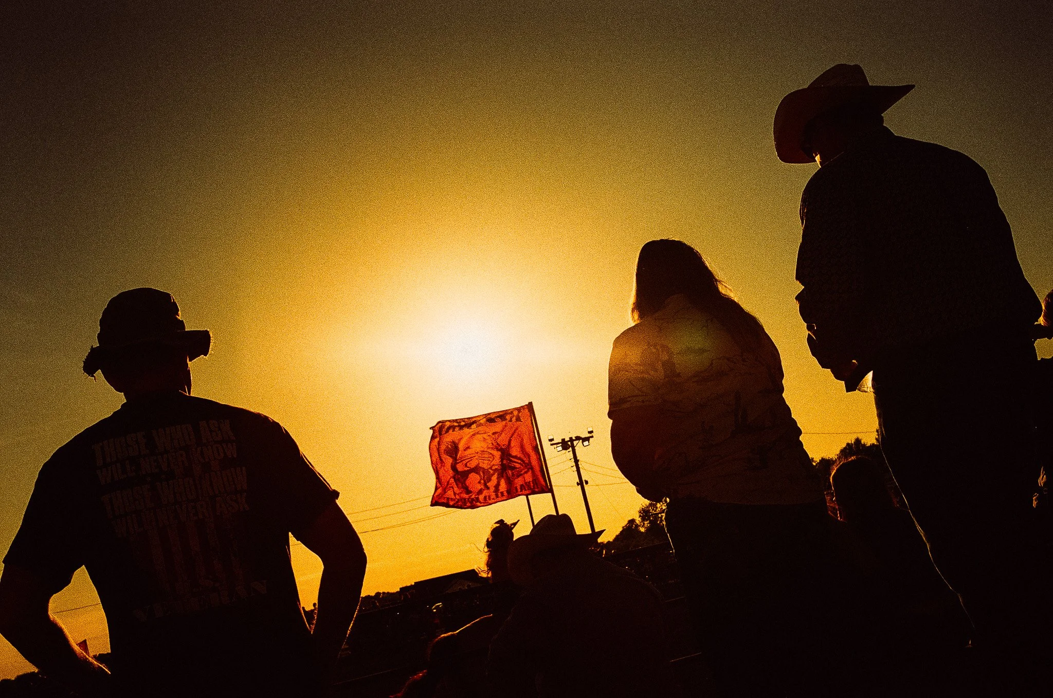 Silhouettes of four people with cowboy hats and a flag against a setting sun.
