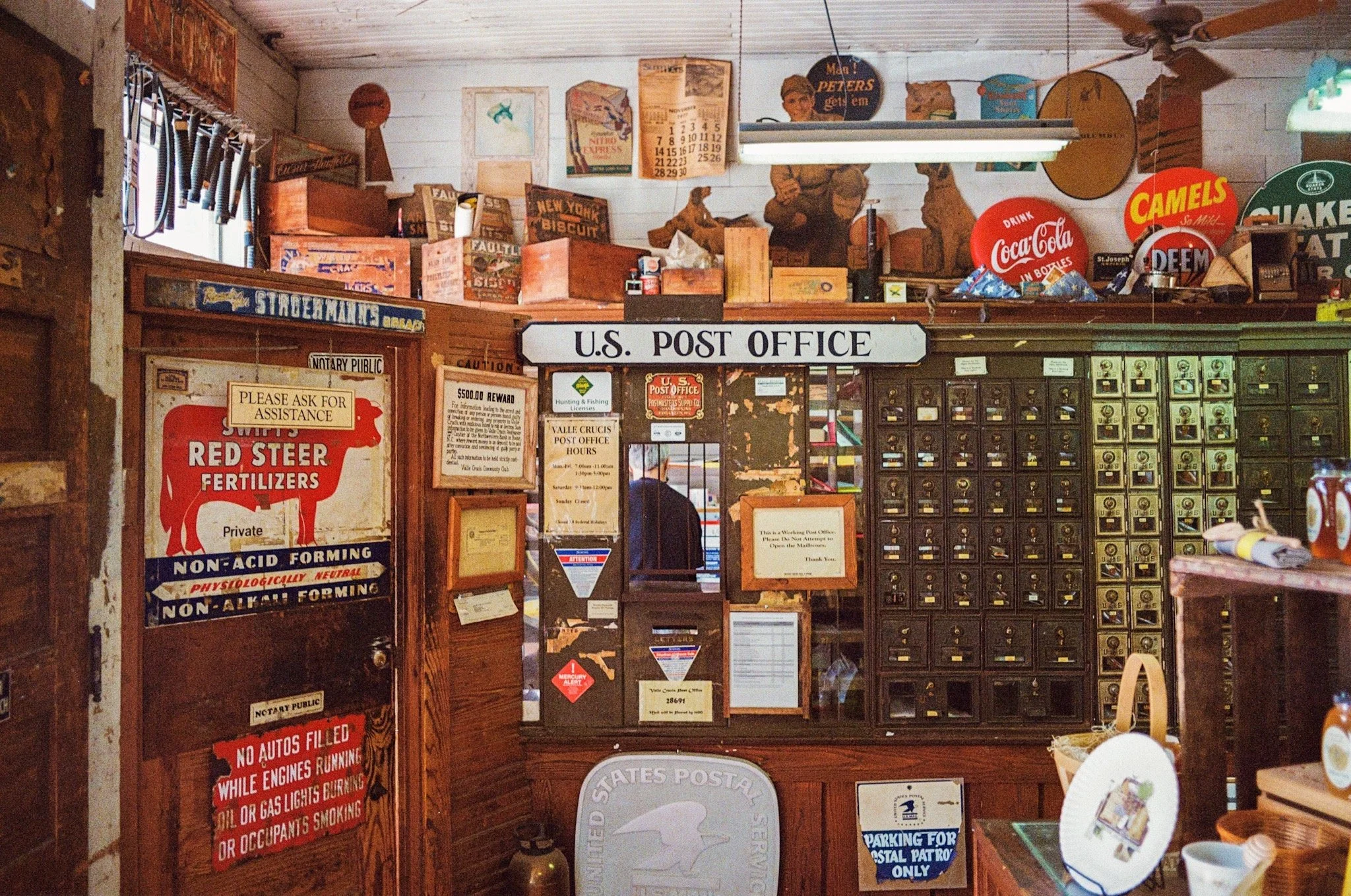Interior of a U.S. Post Office with wooden mailboxes and vintage ads, signs, and collectible items displayed on the walls and shelves.