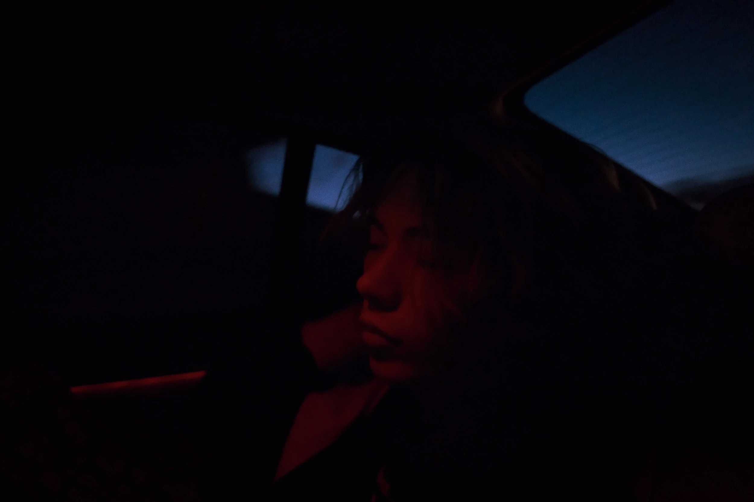 A person with long hair, in a dark setting with minimal lighting and blue hues visible through the window.