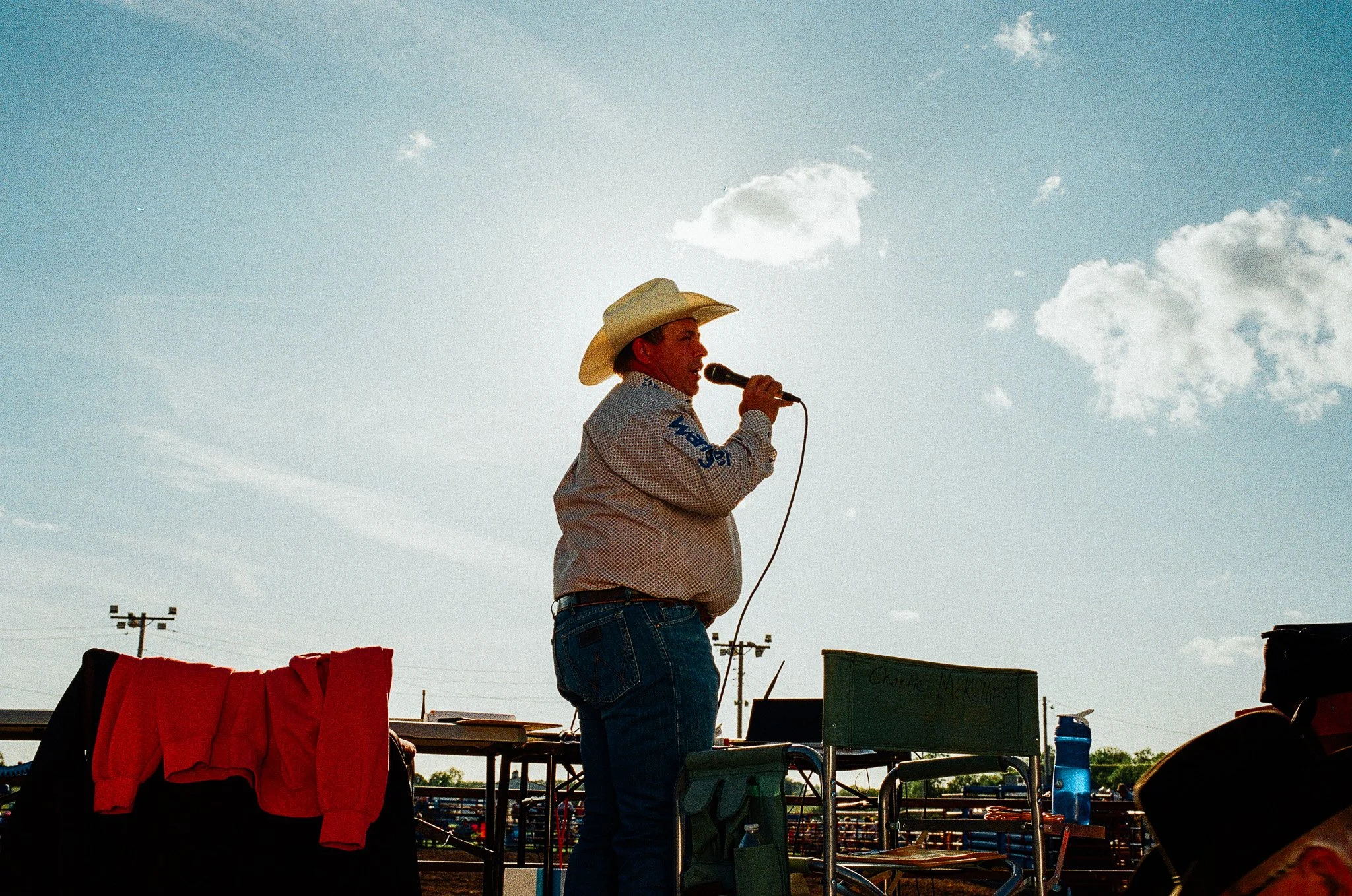 A man wearing a cowboy hat, plaid shirt, and jeans speaking into a microphone at an outdoor event during daytime, with a bright sky and scattered clouds in the background.