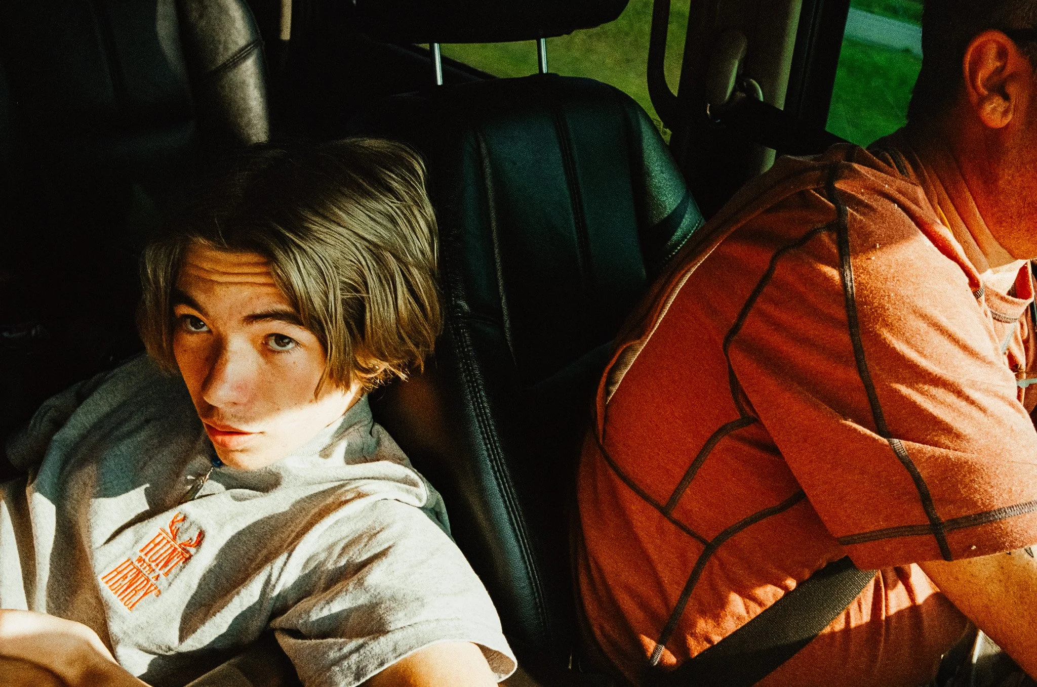 A young man with medium-length blonde hair taking a selfie inside a vehicle, sitting behind the driver, who is wearing a red shirt and facing away.