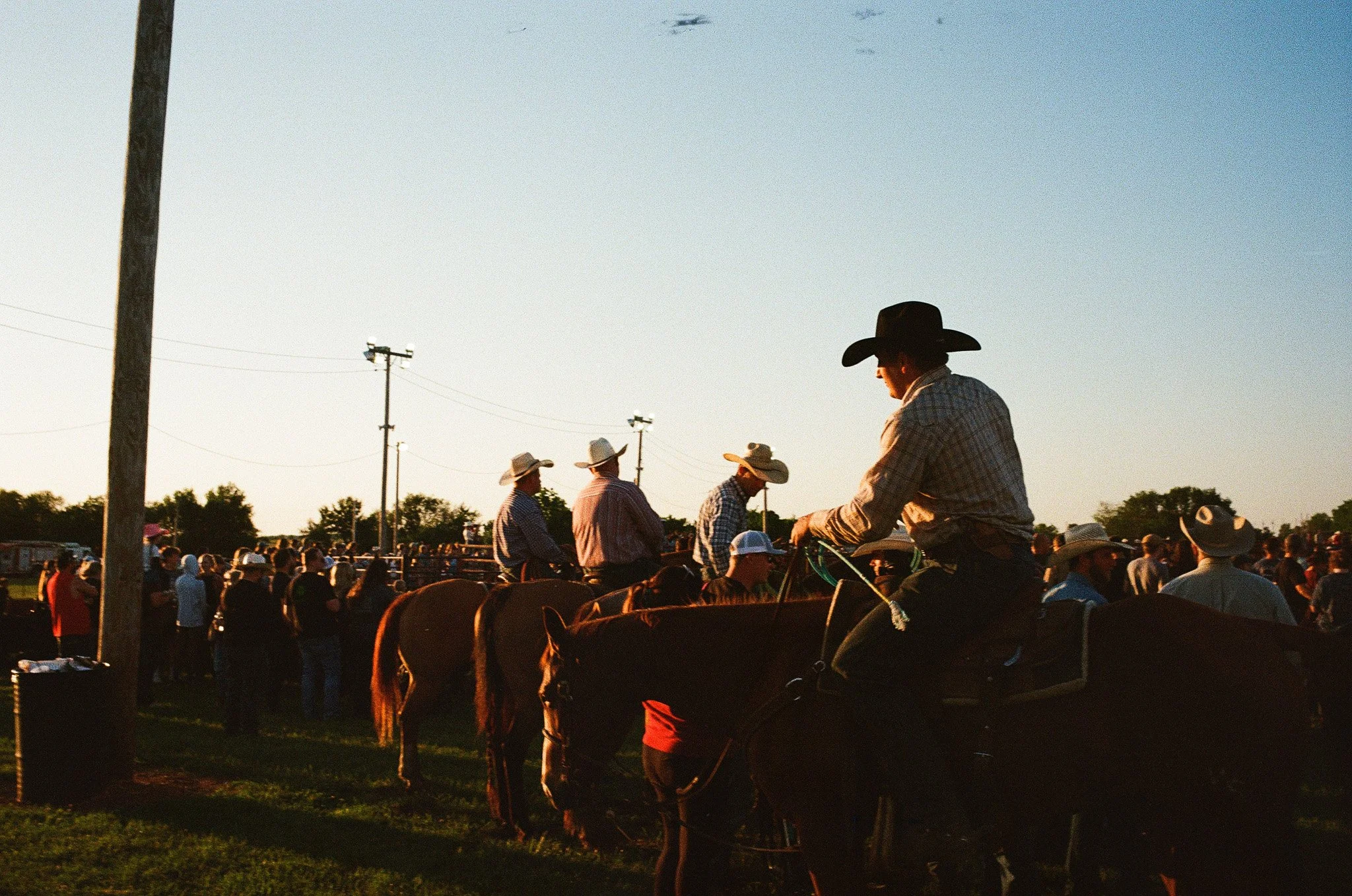 A cowboy in a checkered shirt and black hat sitting on a horse at a rodeo or outdoor event during sunset, with a crowd of people and other cowboys in the background.