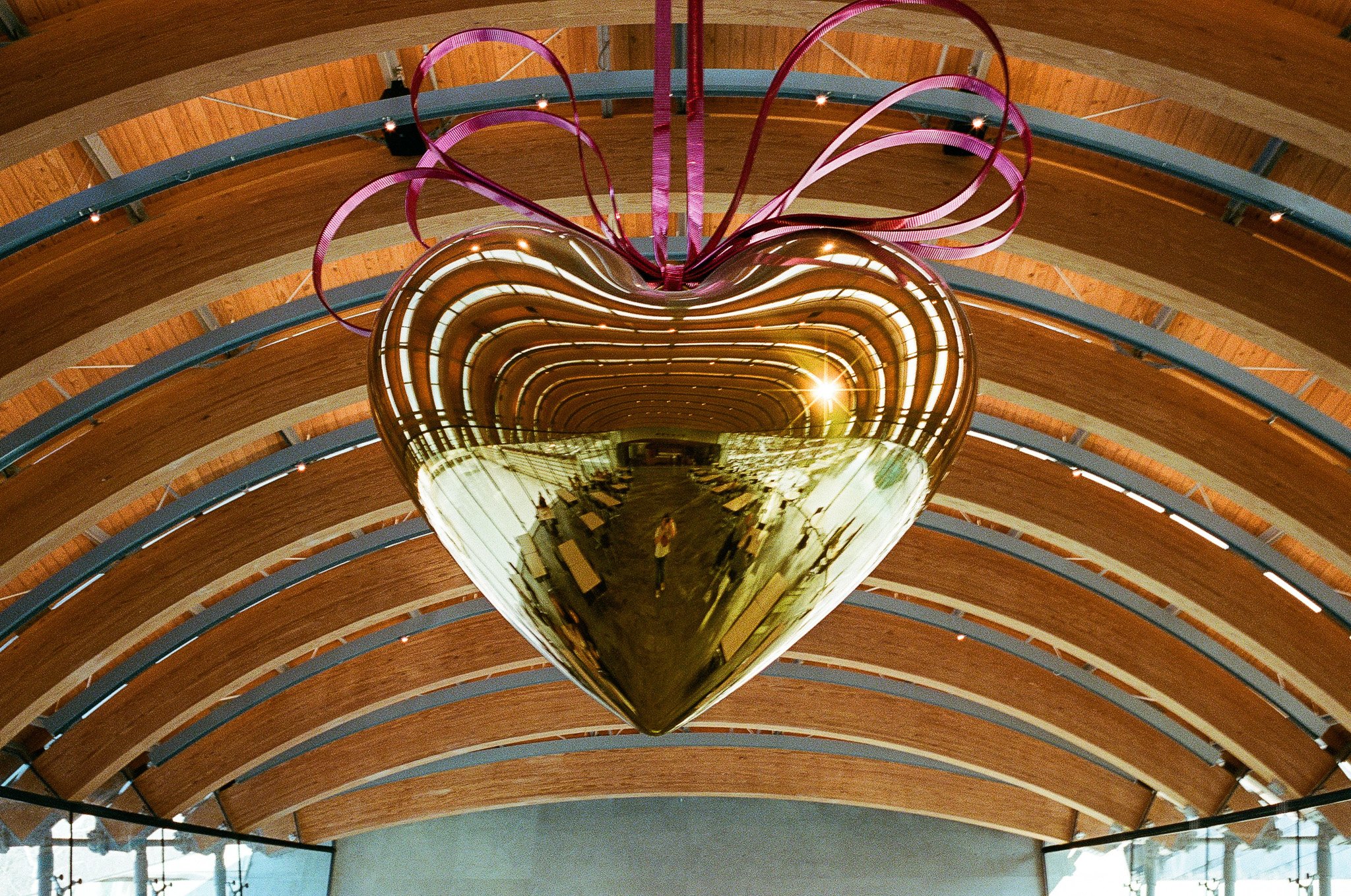 A large, reflective gold heart sculpture hanging from a wooden arched ceiling, adorned with pink ribbon-like ribbons on top.