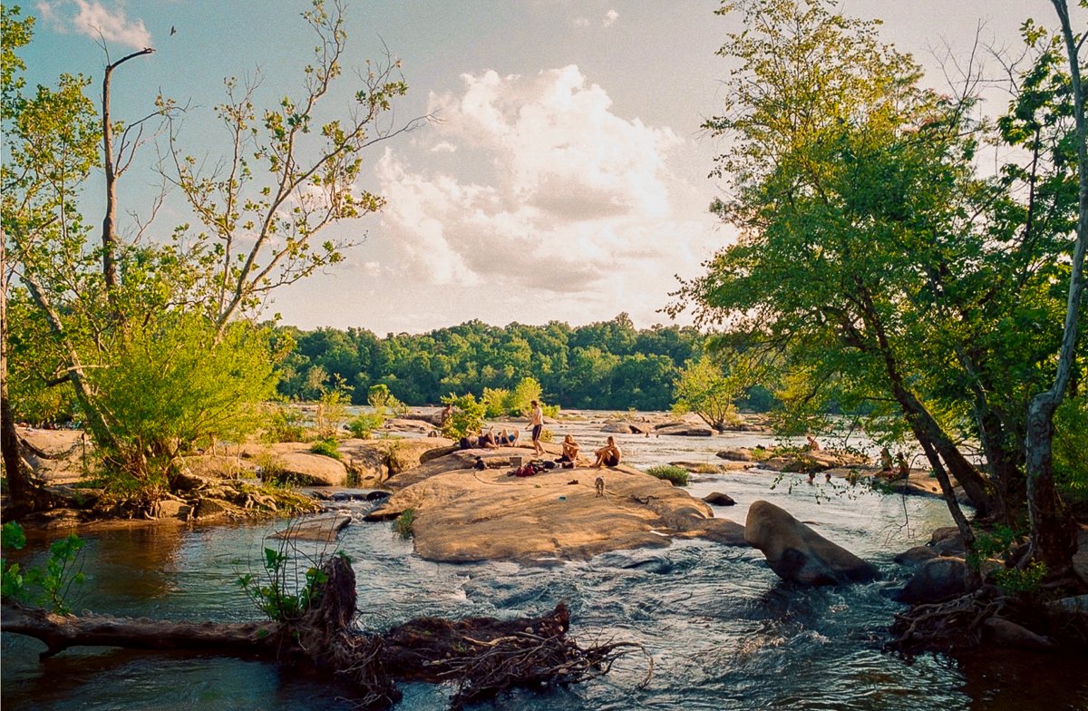 People relaxing on a rocky riverbank surrounded by trees with flowing water nearby