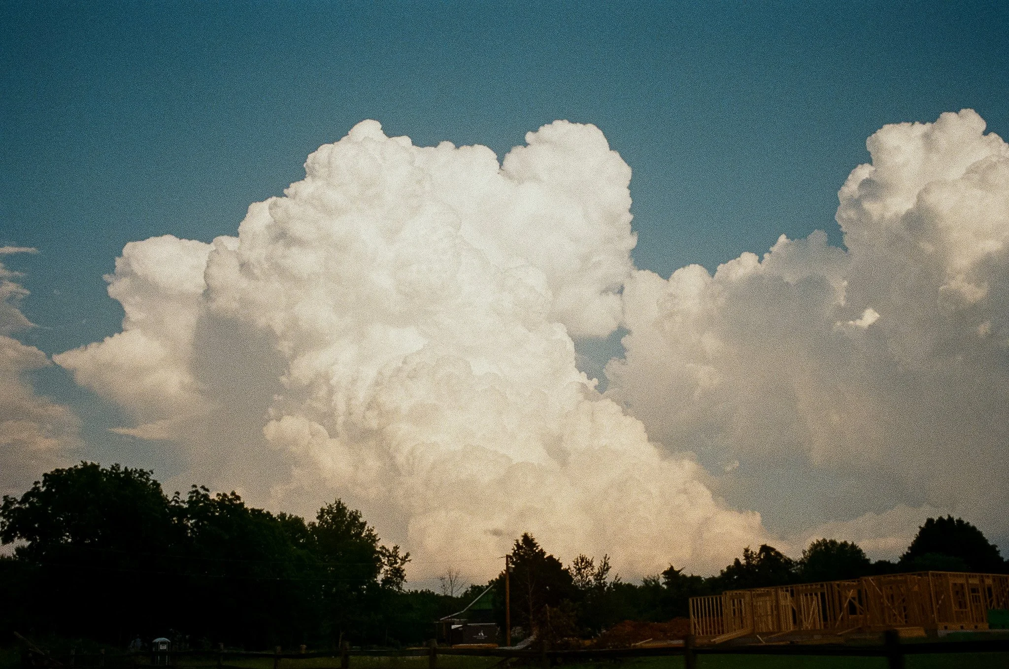 Large white cumulus clouds in the sky over a rural landscape with trees, a house, and a construction site.