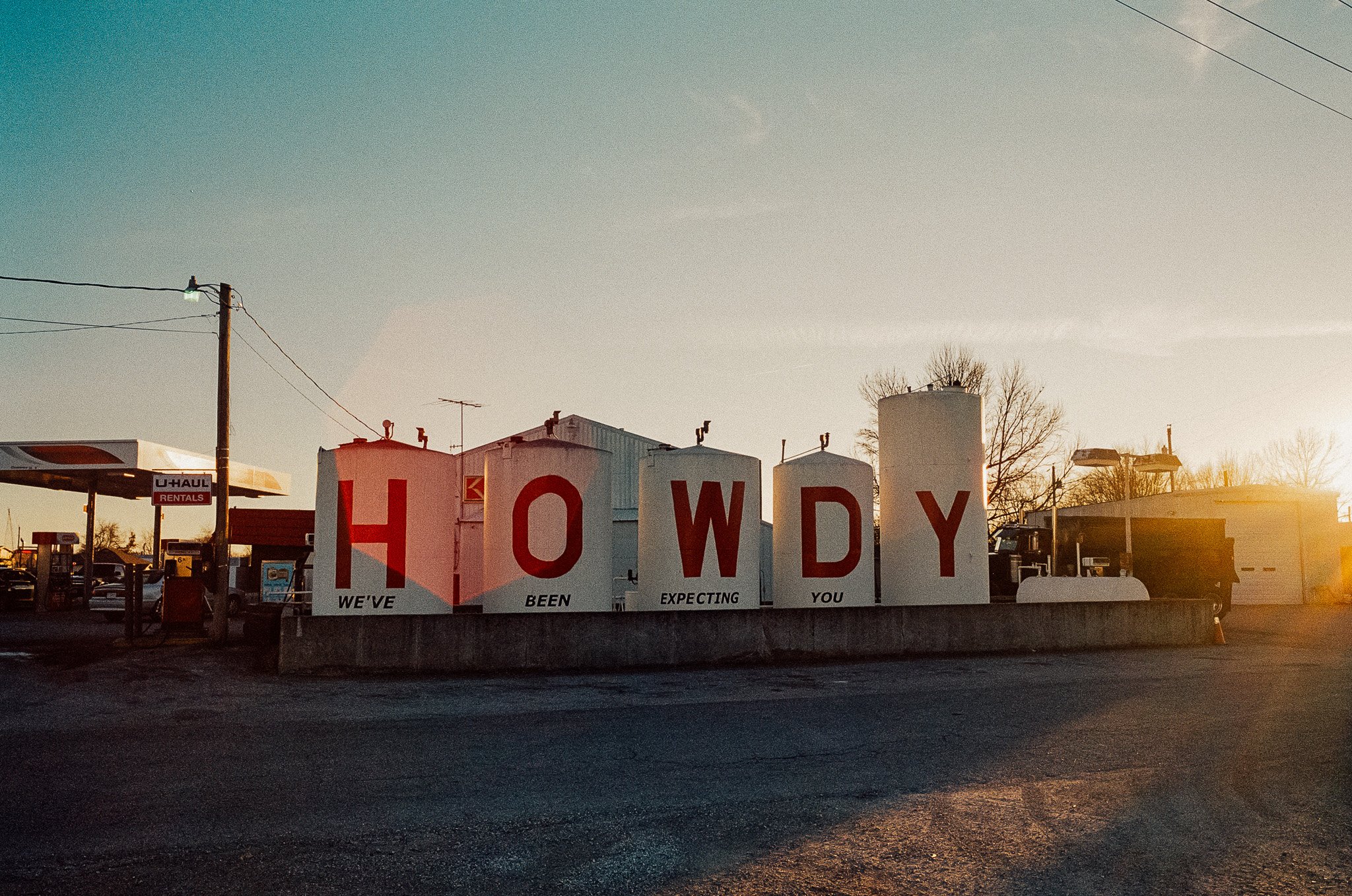 Large roadside sign spelling 'HOWDY' with the words 'WE'VE BEEN EXPECTING YOU' underneath, set against a sunset sky with a parking lot and businesses in the background.