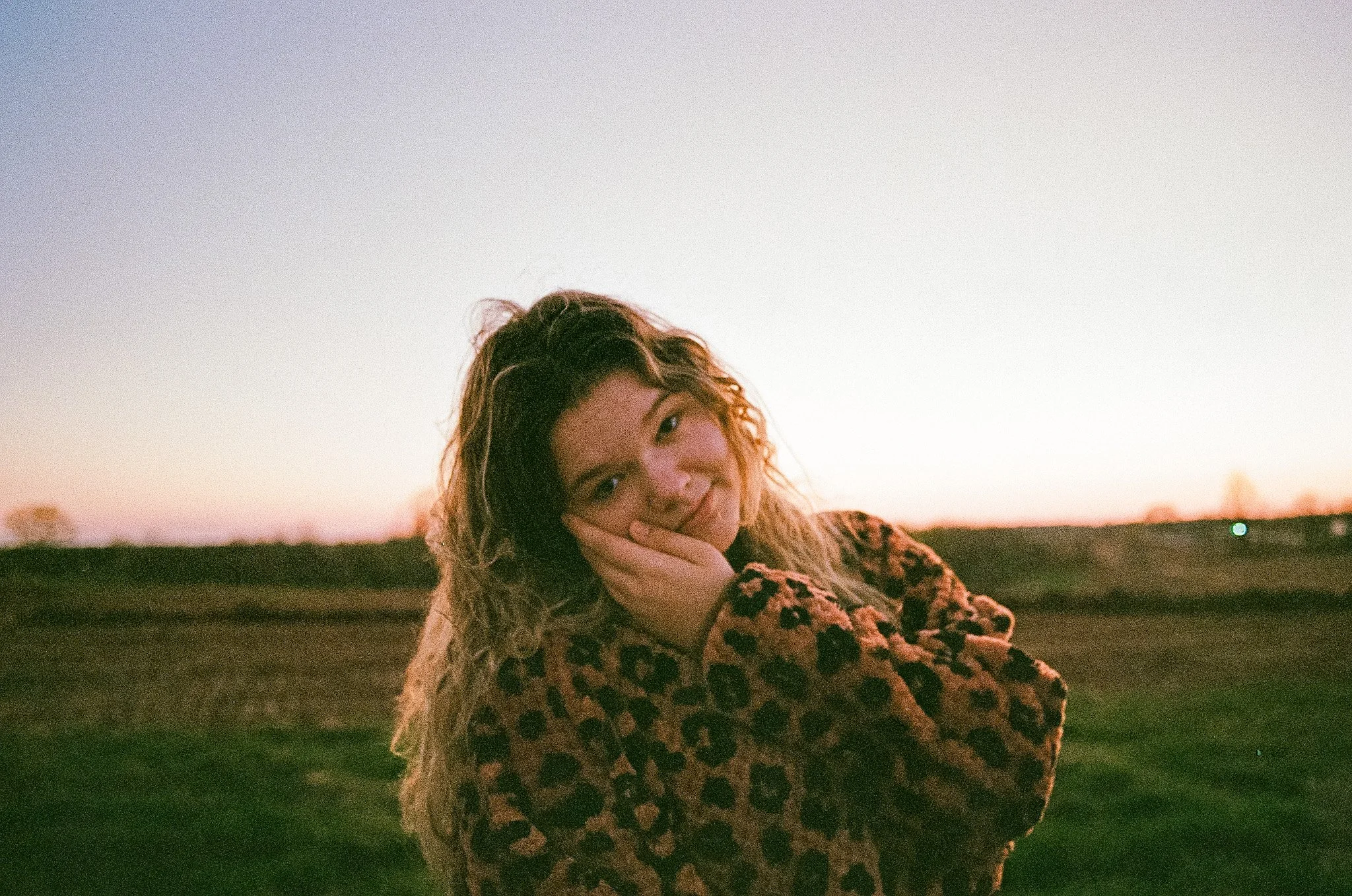 A woman with curly hair wearing a leopard print coat, standing outdoors during sunset with a scenic landscape in the background.