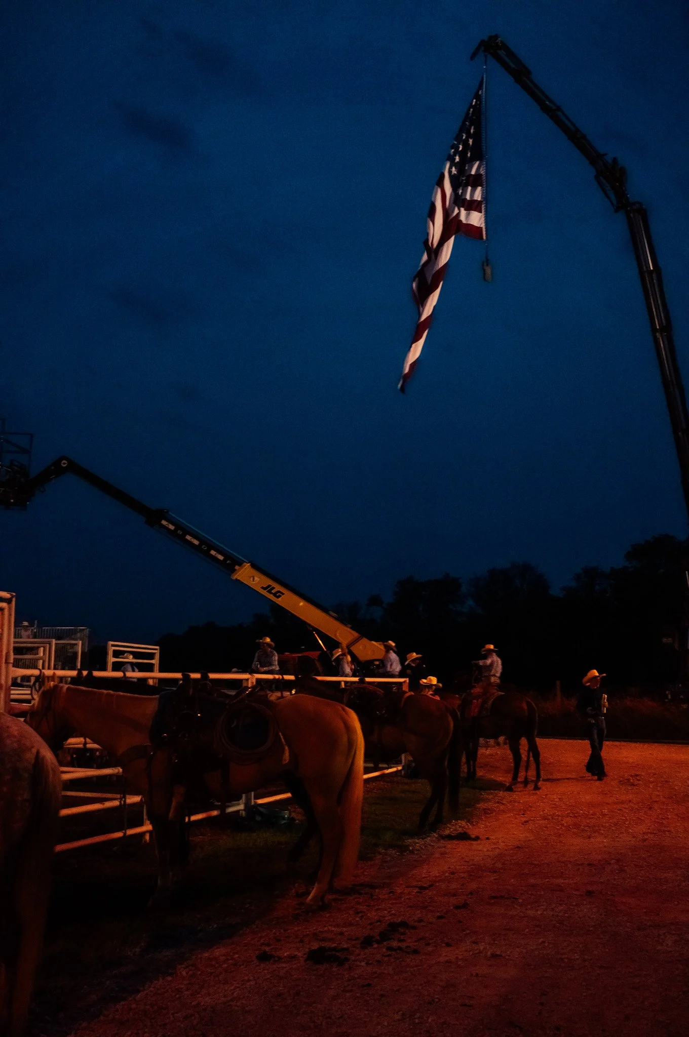 Nighttime scene at a rodeo or western event with mounted cowboys, a large American flag hanging from a crane, and a dark blue sky.