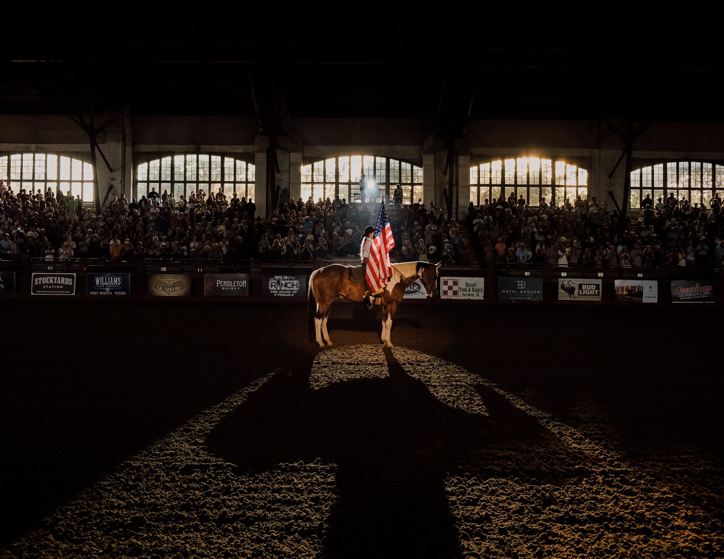 A person riding a horse in an indoor arena, holding an American flag, with a large crowd seated in the background and sunlight illuminating through windows.