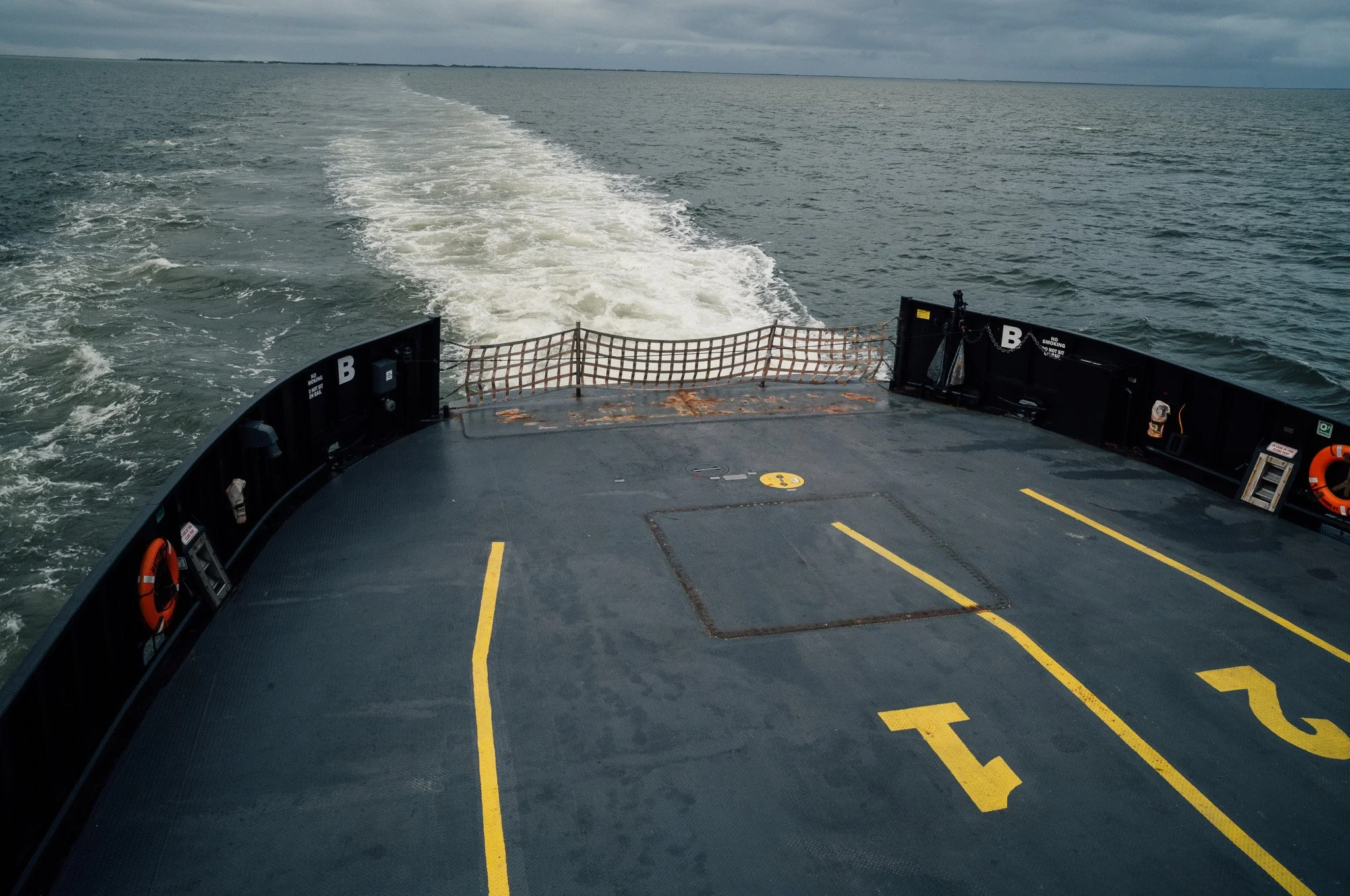View of the deck of a ship moving through the ocean, with water and sky visible. The deck has yellow markings and safety equipment, and the wake trails behind the ship.