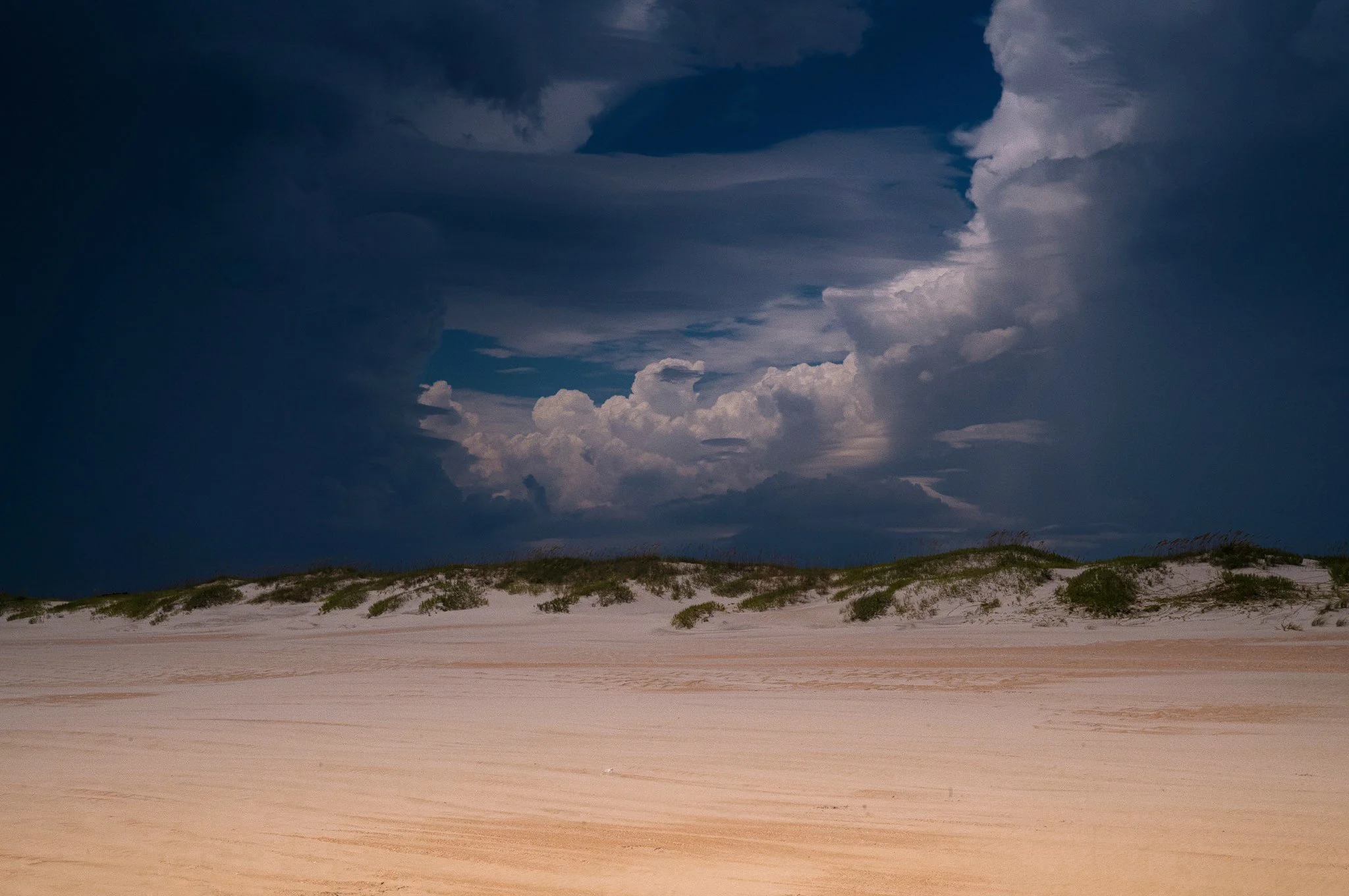A deserted sandy beach with dunes and sparse vegetation, under a dark, cloudy sky.