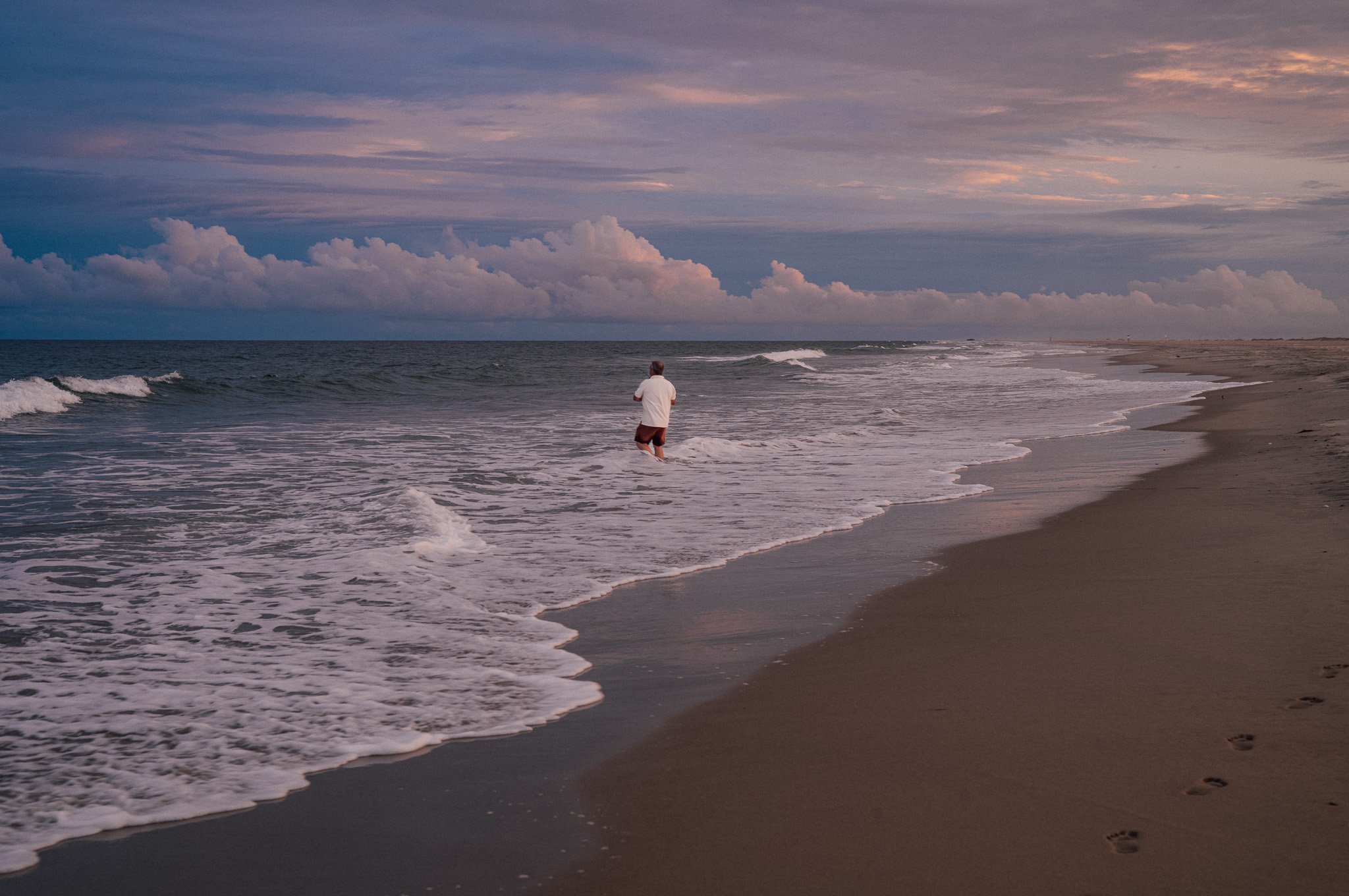 A person walking along the shoreline of a beach at sunset, with footprints in the sand and waves washing up on the shore.