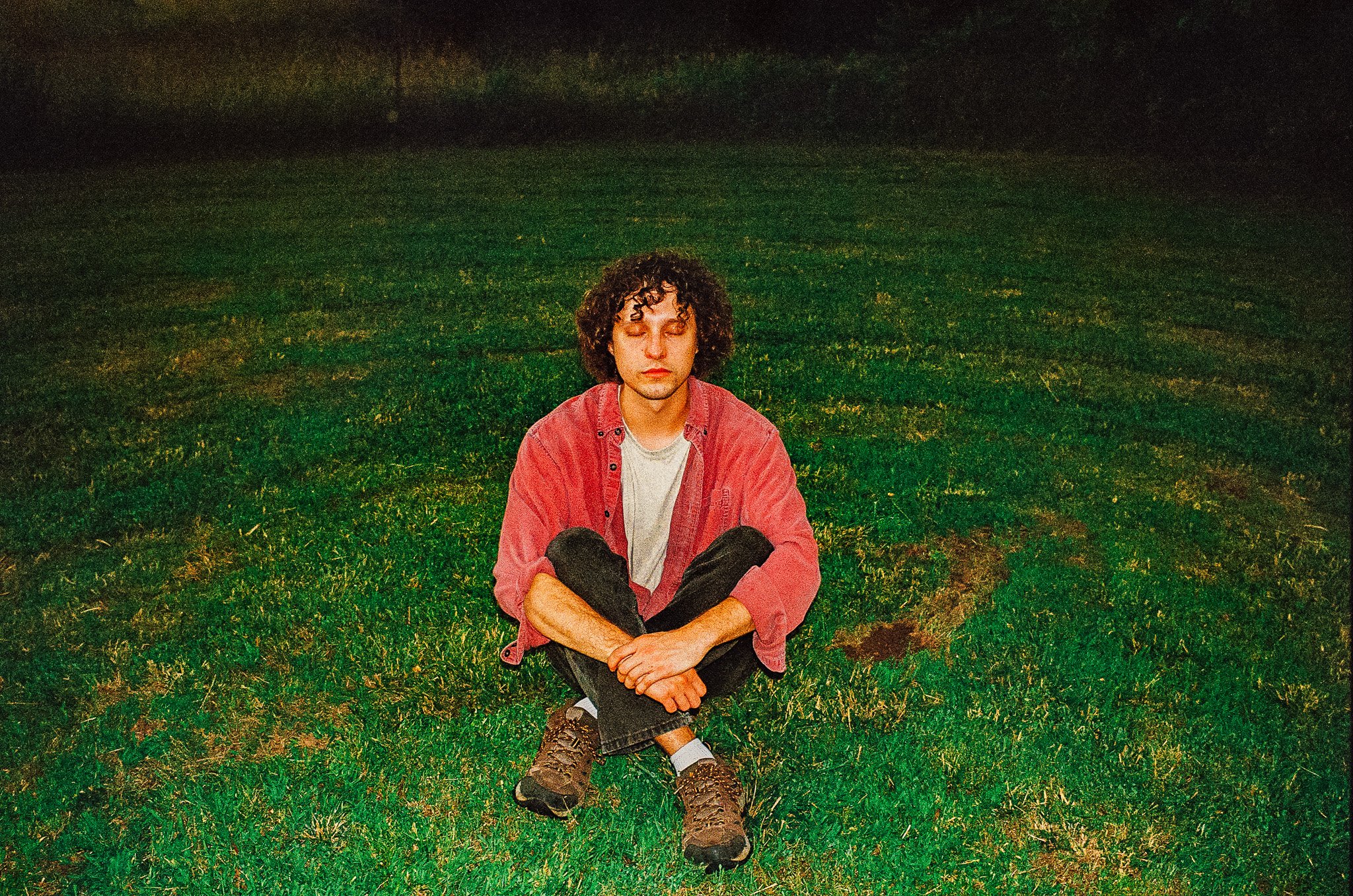 A young man with curly hair sitting cross-legged on a grassy field at night, eyes closed, wearing a red jacket, gray t-shirt, black pants, and hiking shoes.