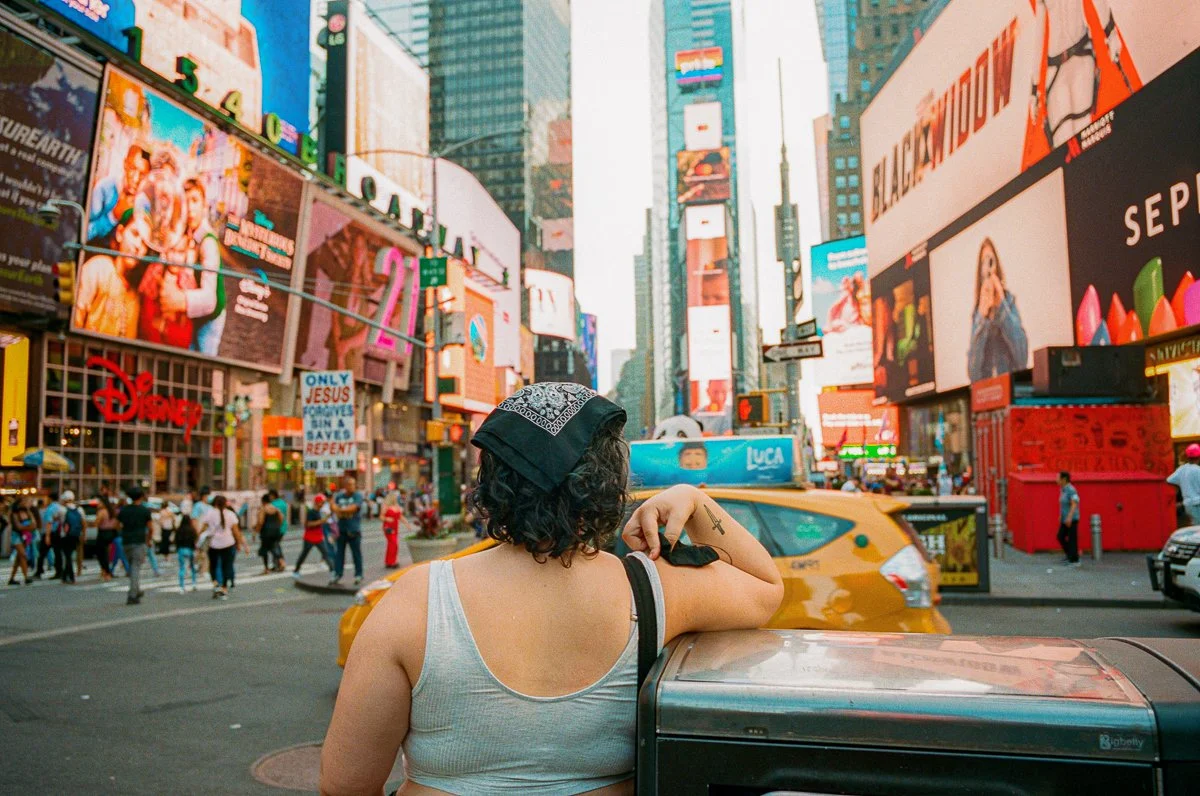 Person with curly hair wearing a bandana and a gray tank top leaning on a mailbox in Times Square, New York City, surrounded by bright digital billboards and taxis.