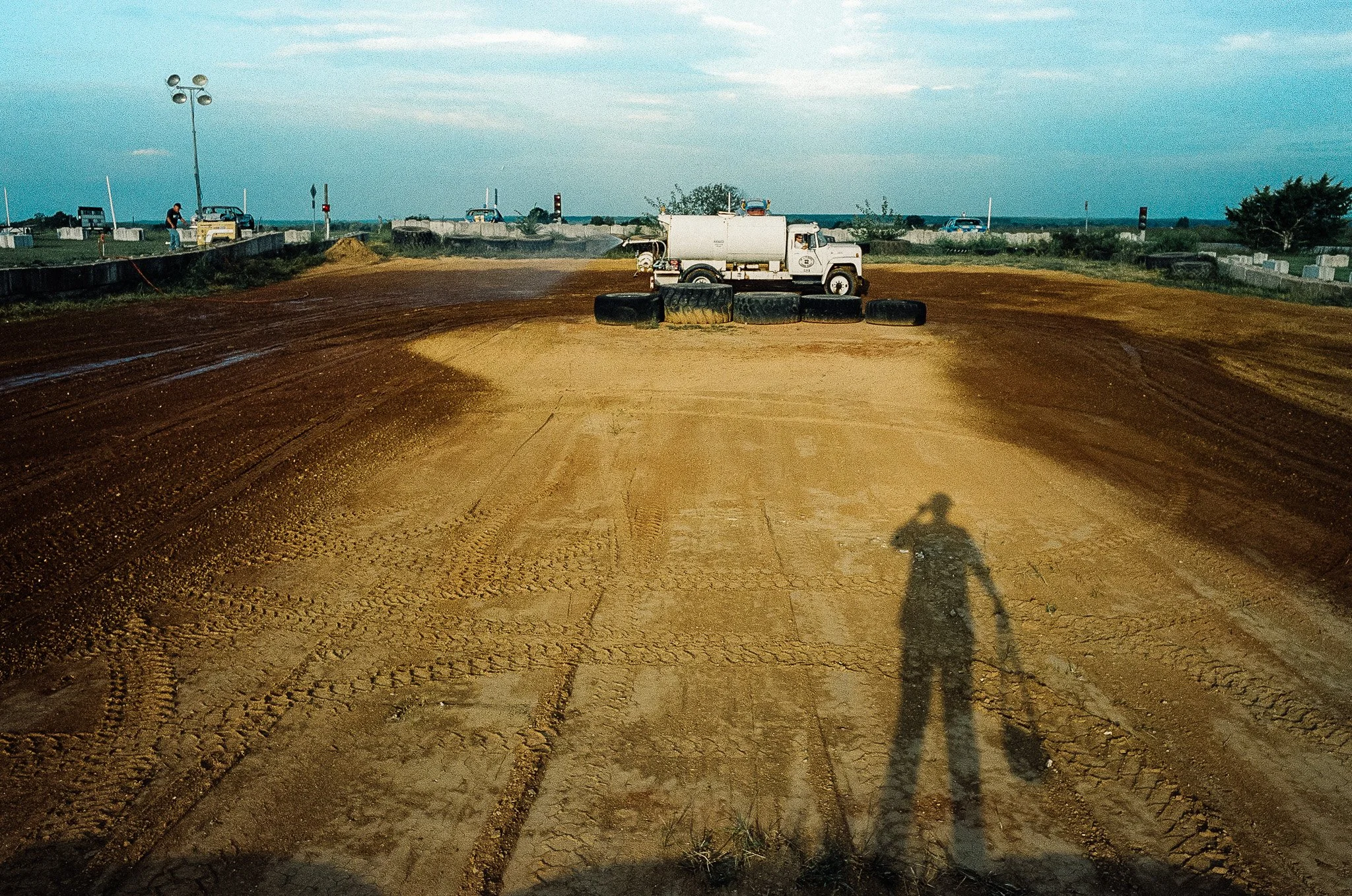 A dirt race track with a white truck and spare tires in the center, and a person’s shadow taking the photo in the foreground. There are cars and a person working near the track in the background under an overcast sky.