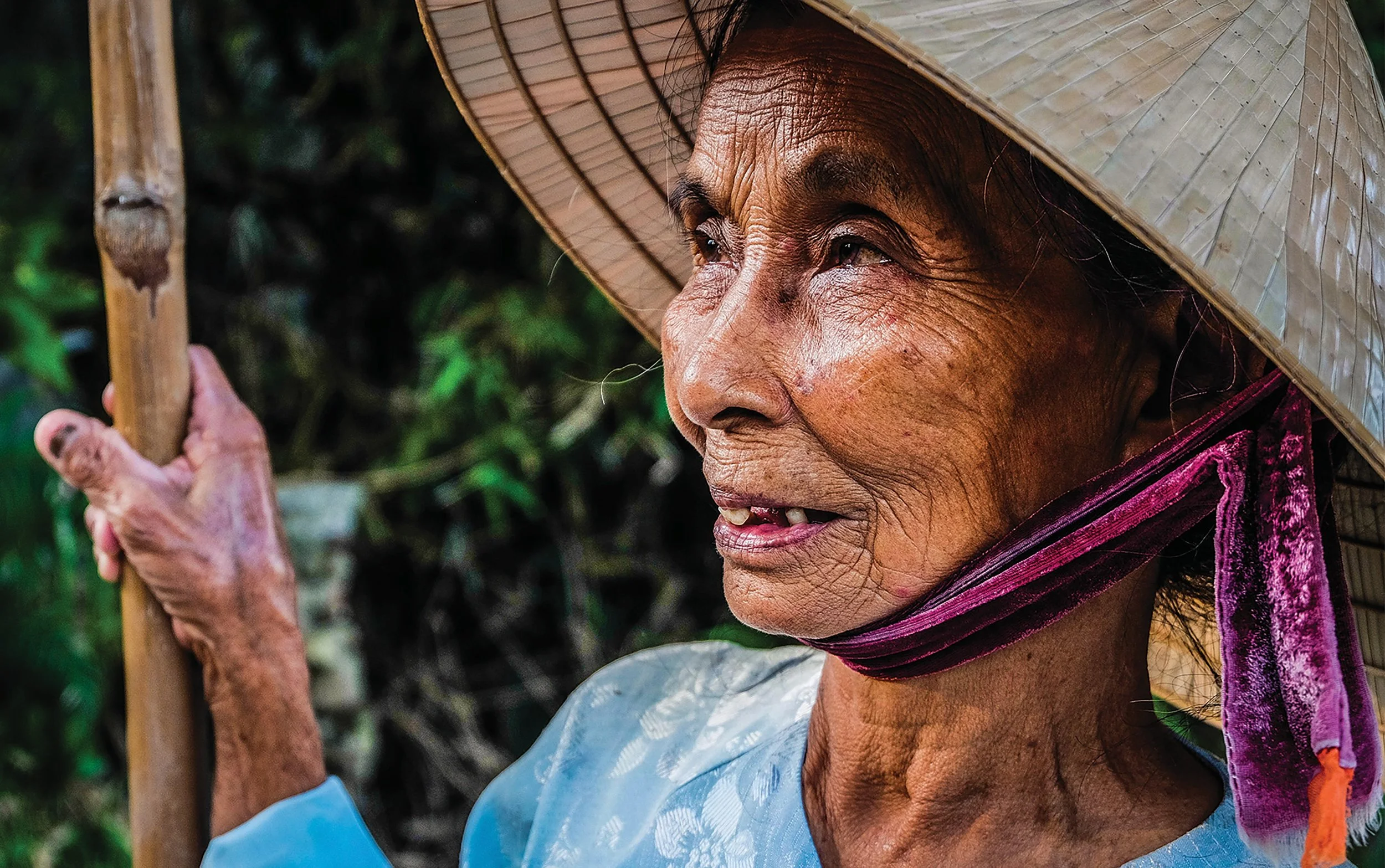 Close-up of an elderly woman with a weathered face, wearing a wide-brimmed straw hat and a purple cloth face mask hanging below her chin, holding a wooden stick, outdoors with greenery in the background.