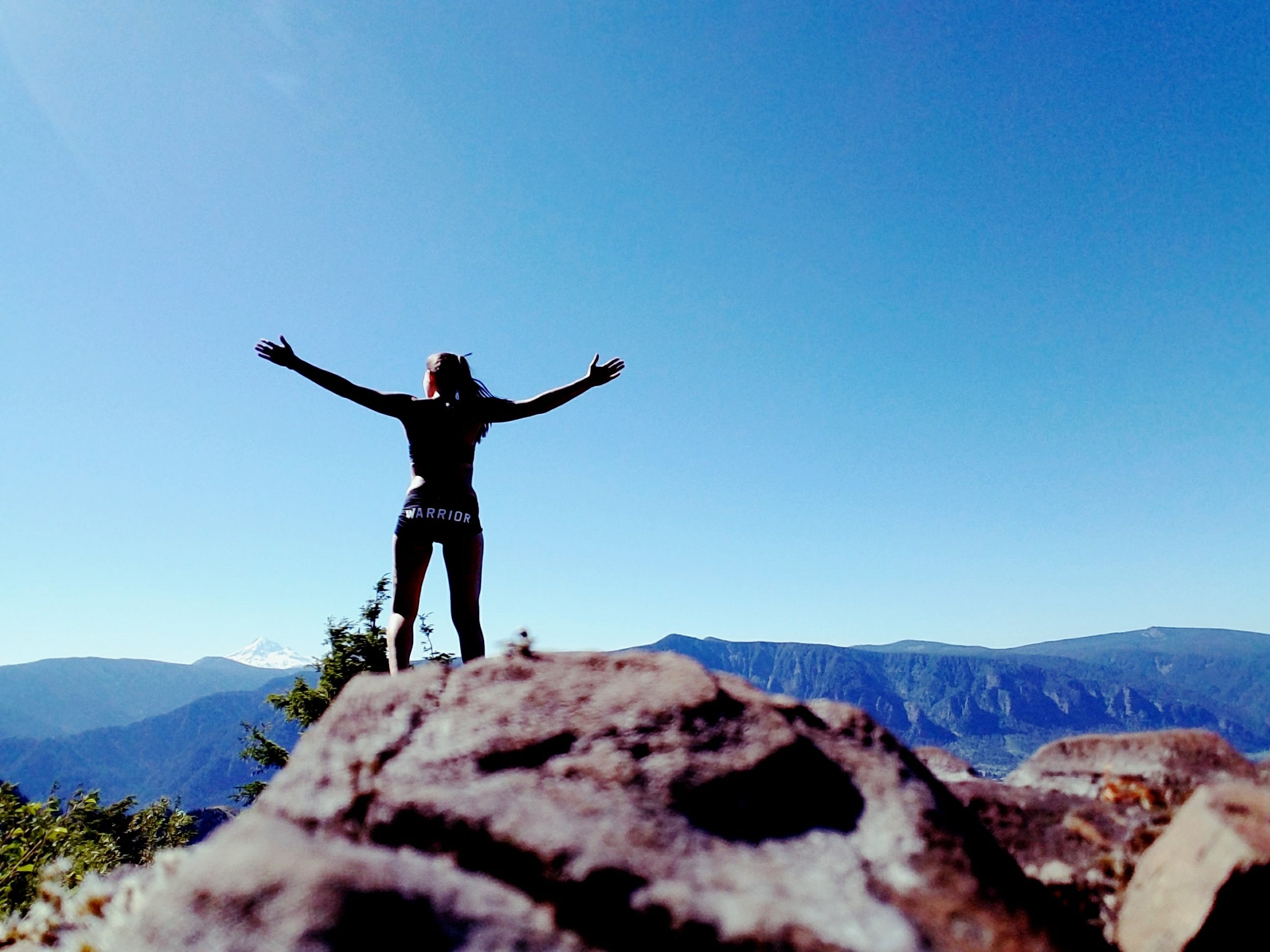 A person standing on a rock with arms outstretched, overlooking a mountain landscape under a clear blue sky.