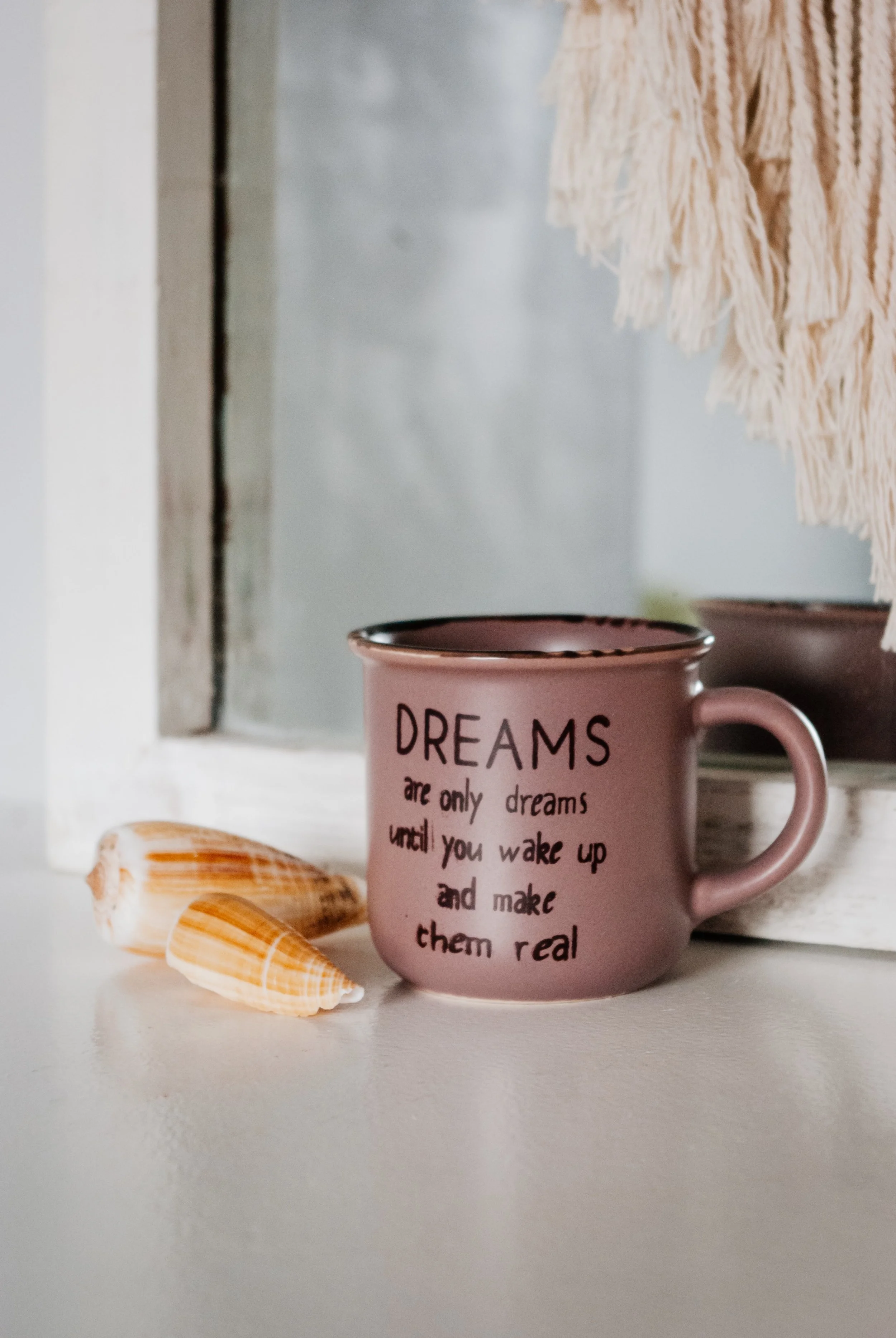A pink mug with black text saying "DREAMS are only dreams until you wake up and make them real" on a white surface, with two seashells beside it and a fringed cloth hanging in the background.