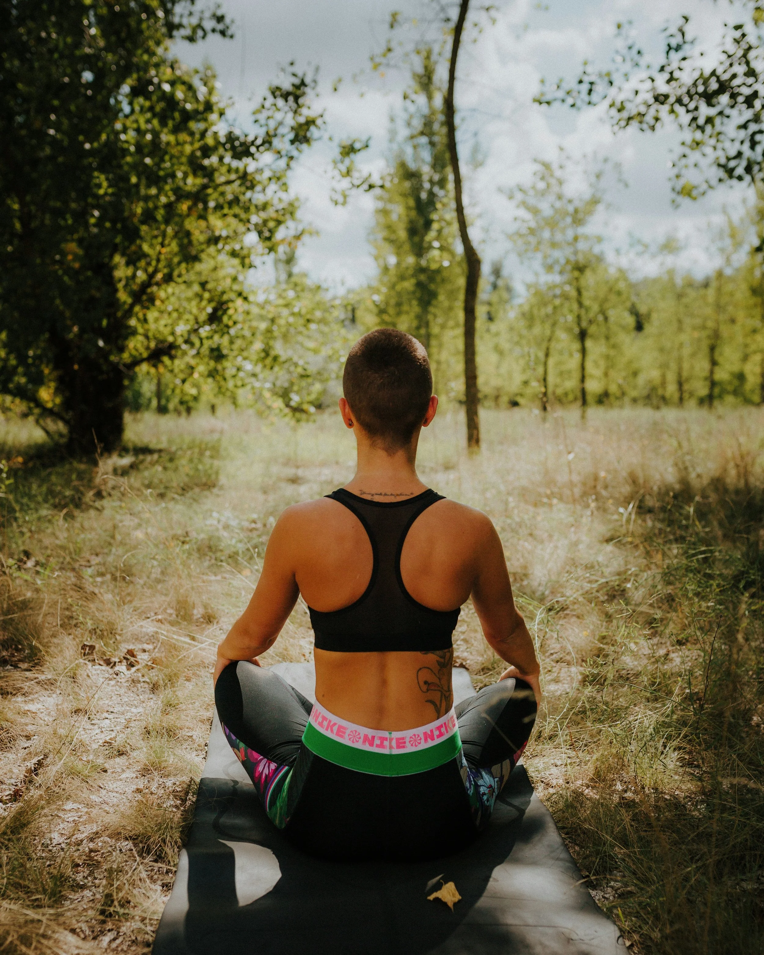 Person practicing yoga outdoors in a forested area, sitting cross-legged on a yoga mat facing away from the camera.