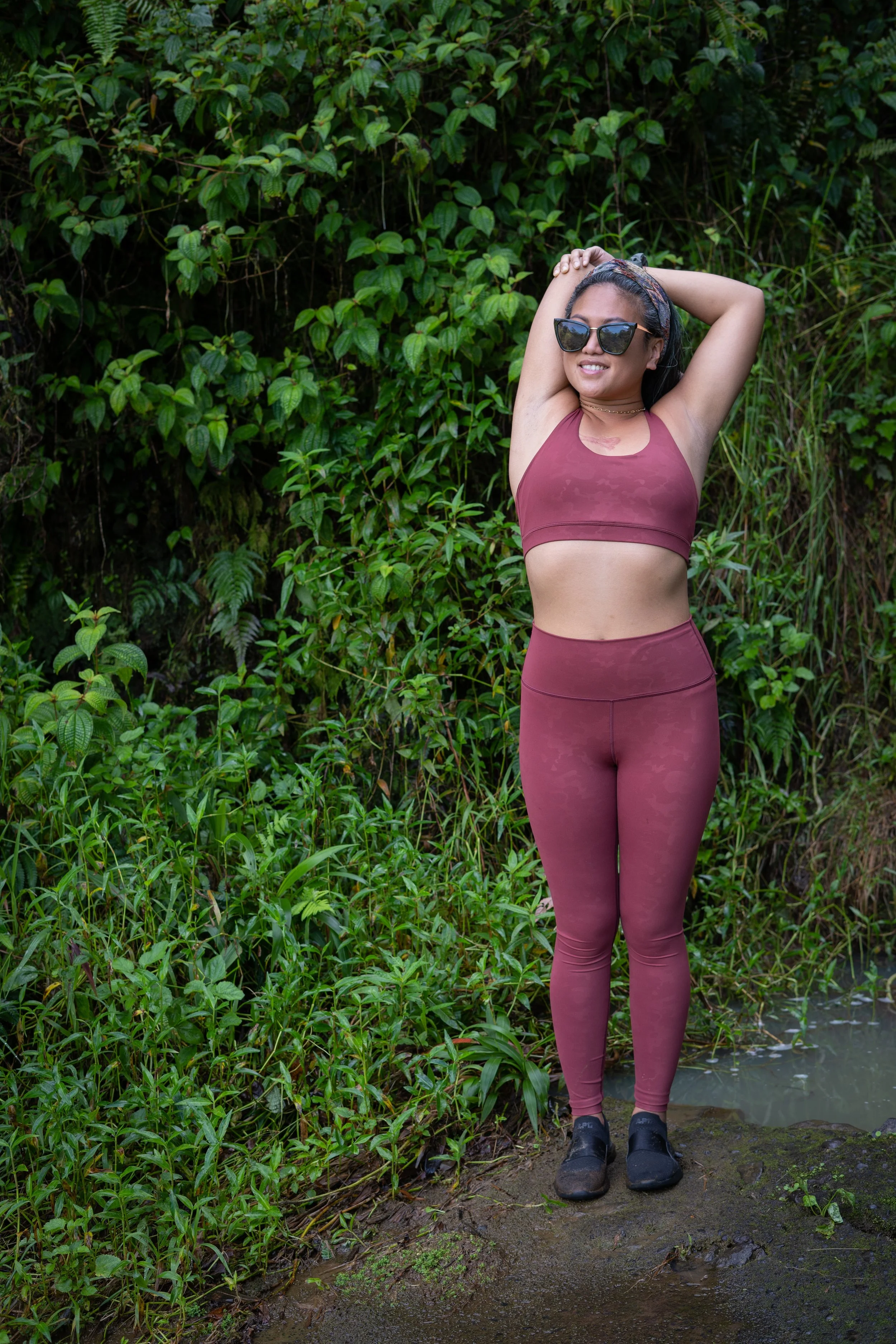 A woman wearing sunglasses and activewear standing in a lush green outdoor setting with her arms raised behind her head.