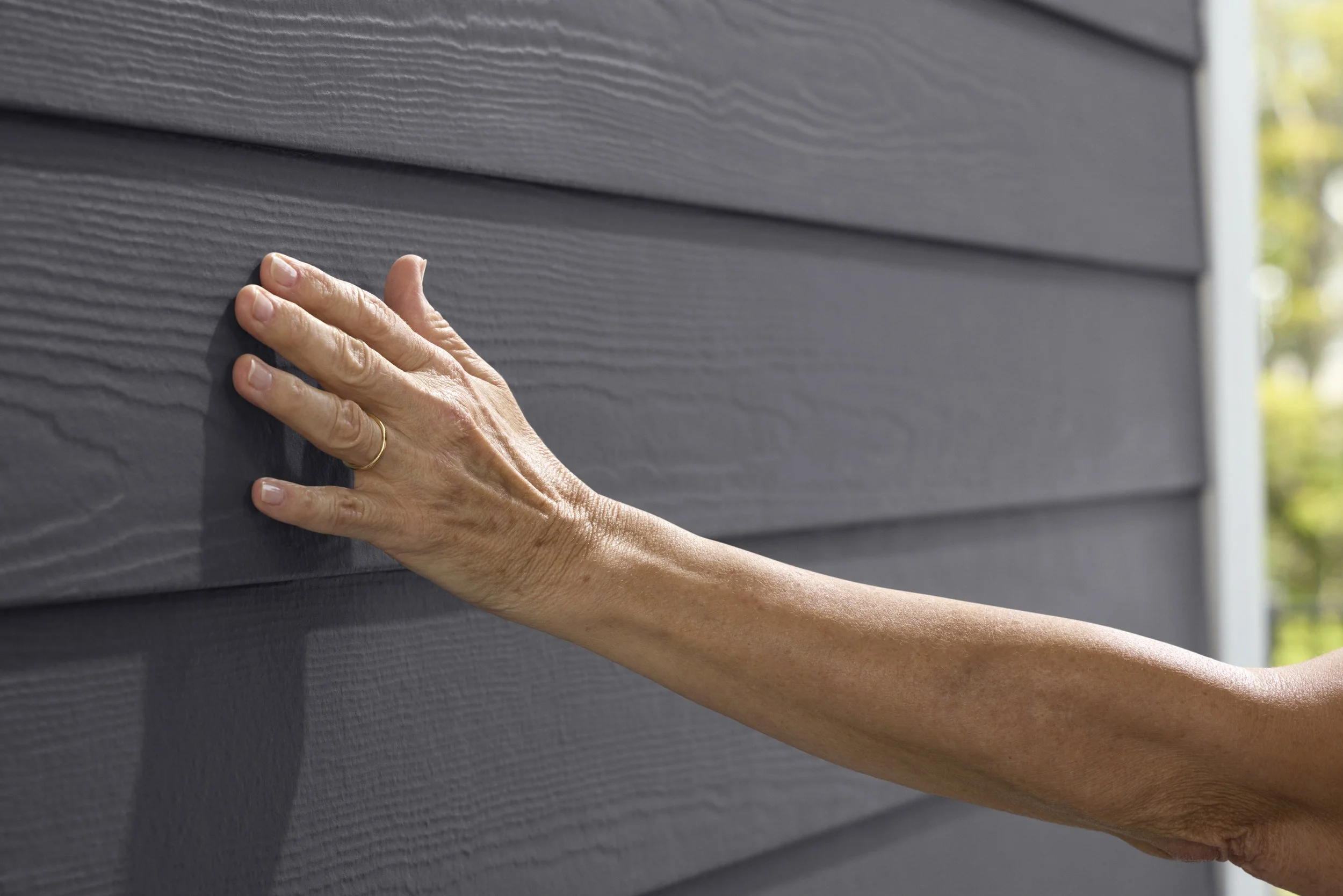 A person's hand touching gray horizontal siding on a building, with blurred background of trees and sunlight.