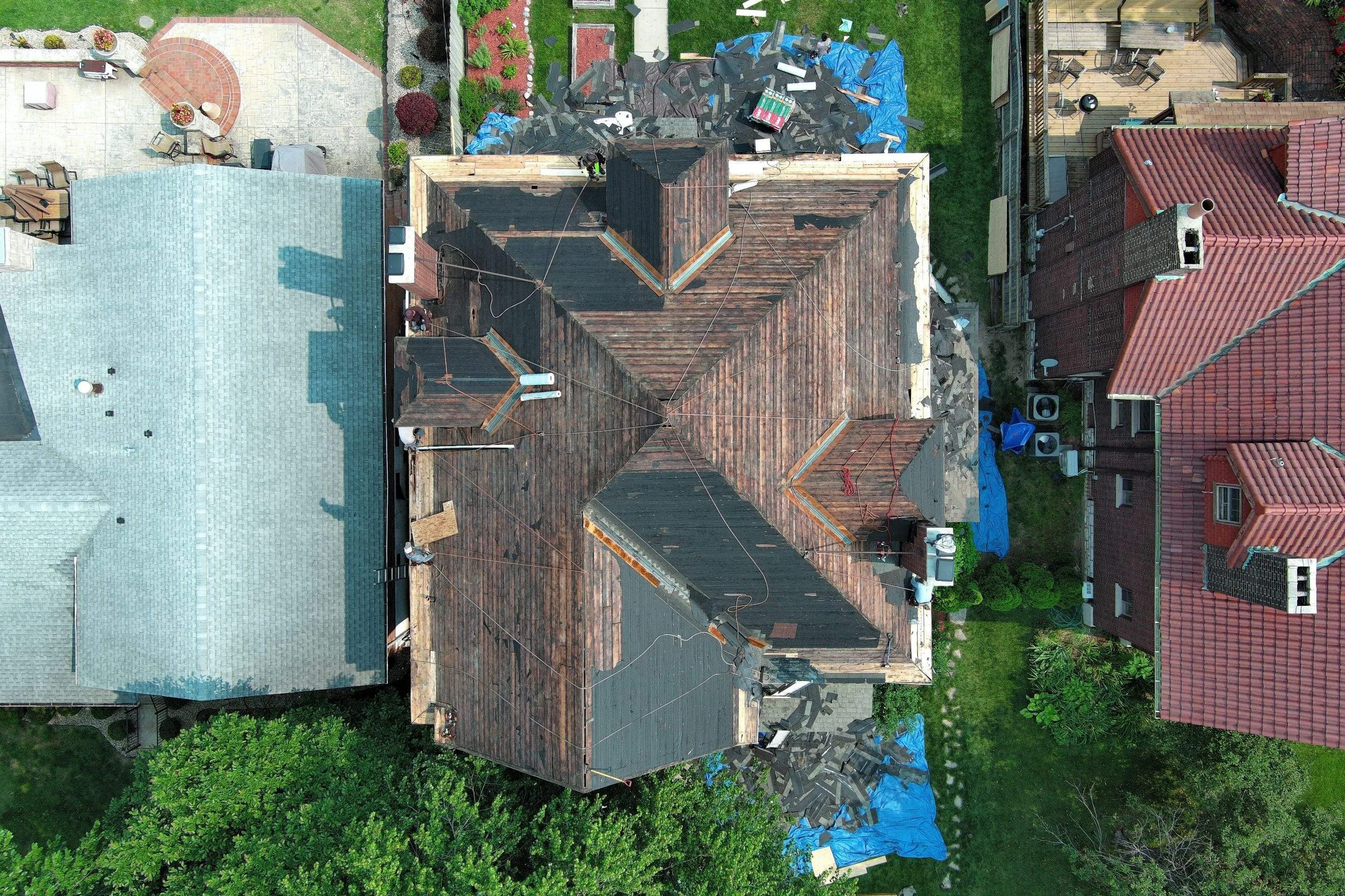An aerial view of a house under renovation with a construction worker on the roof, roofing materials, and surrounding yard with neighboring houses, trees, and plants.
