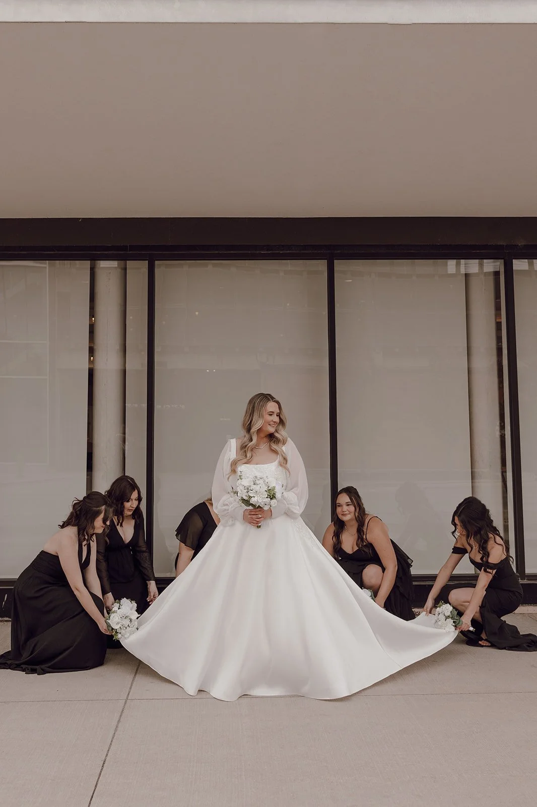 A bride in a white wedding gown holding a bouquet, surrounded by five bridesmaids in black dresses, sitting or kneeling on the sidewalk in front of a large glass window.