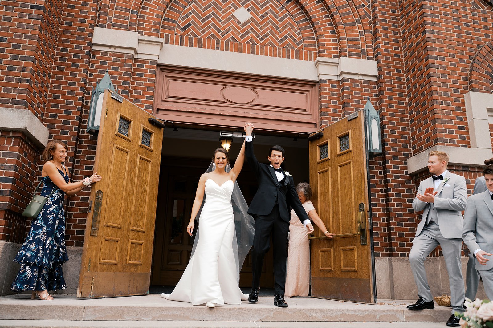 A bride and groom are celebrating their wedding outside a church with open wooden doors, surrounded by well-dressed guests clapping and smiling.