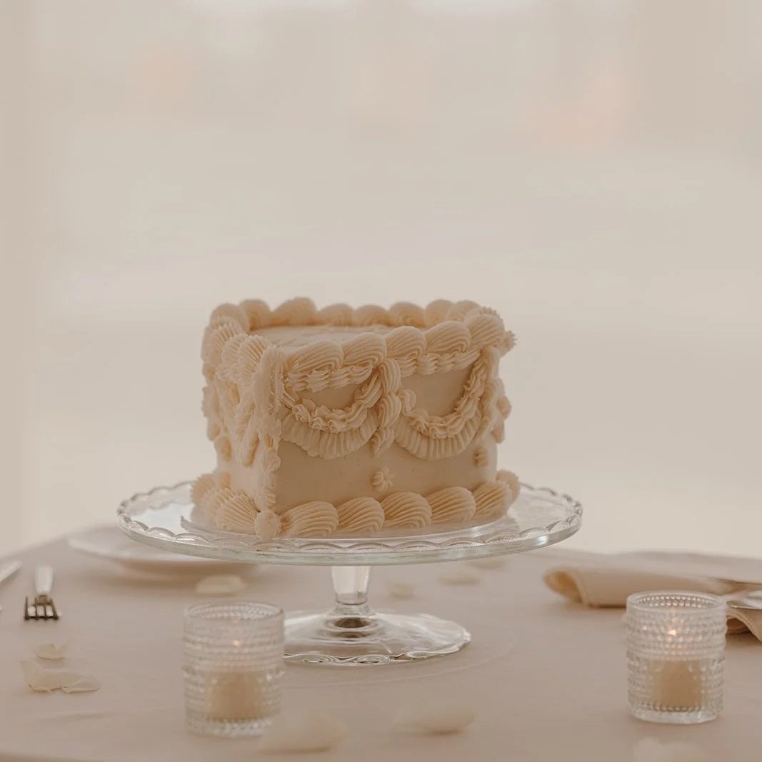 A decorated white cake with piped frosting, placed on a glass cake stand on a white table, with lit candles, a fork, napkins, and a knife nearby.
