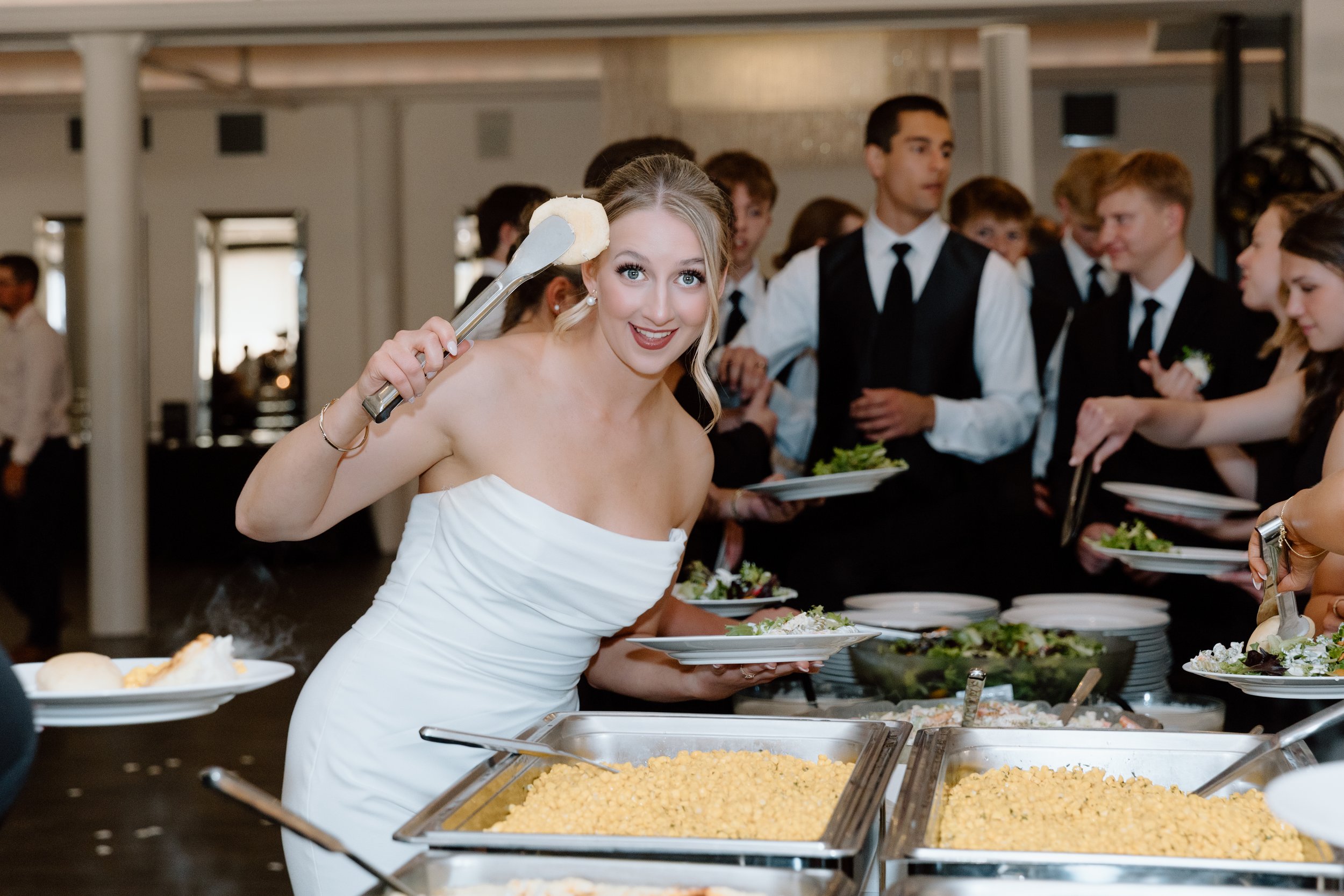 A bride in a white strapless wedding dress serving herself at a buffet table with various dishes, while surrounded by guests at a wedding reception.