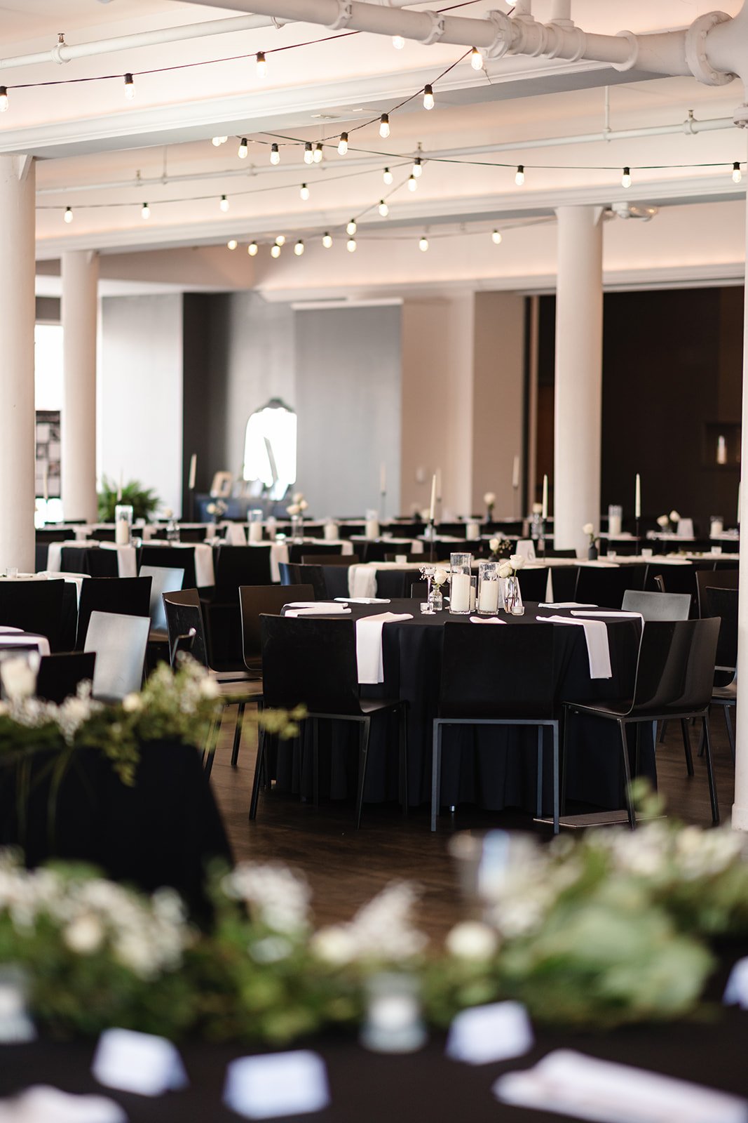 Elegant event space with round tables covered in black tablecloths, white napkins, candles, floral centerpieces, and string lights hanging from the ceiling.