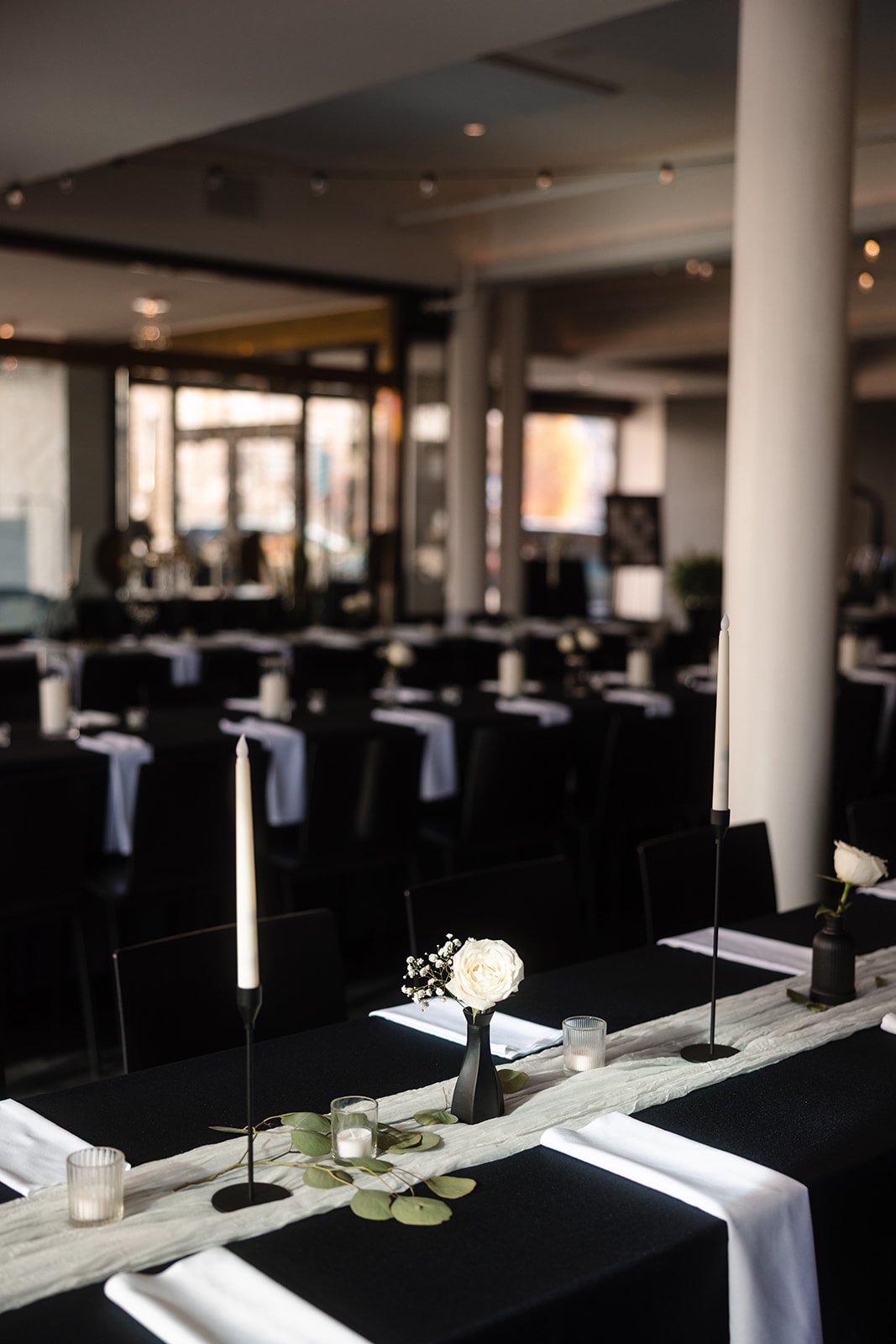 Elegant table setup in a restaurant with tall black candles, white flowers in black vases, and small candles, with black tablecloths and white napkins, in a modern dining space with large windows.