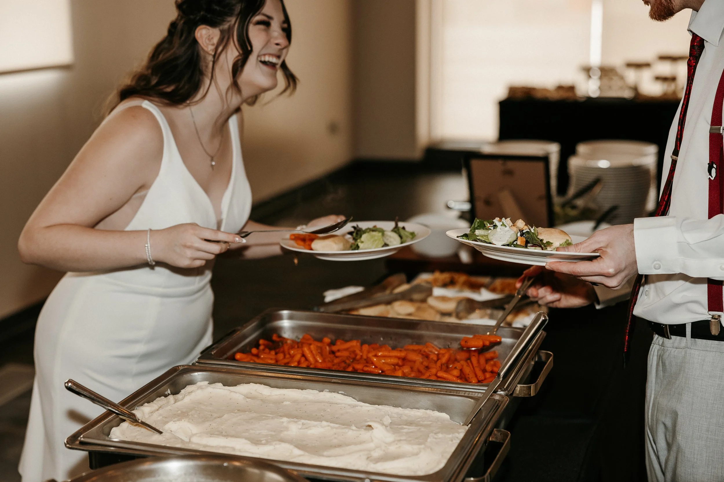 A woman in a white dress laughing while holding a plate of salad at a buffet. A man in a white shirt with suspenders is serving food, including carrots and mashed potatoes, at a buffet table.