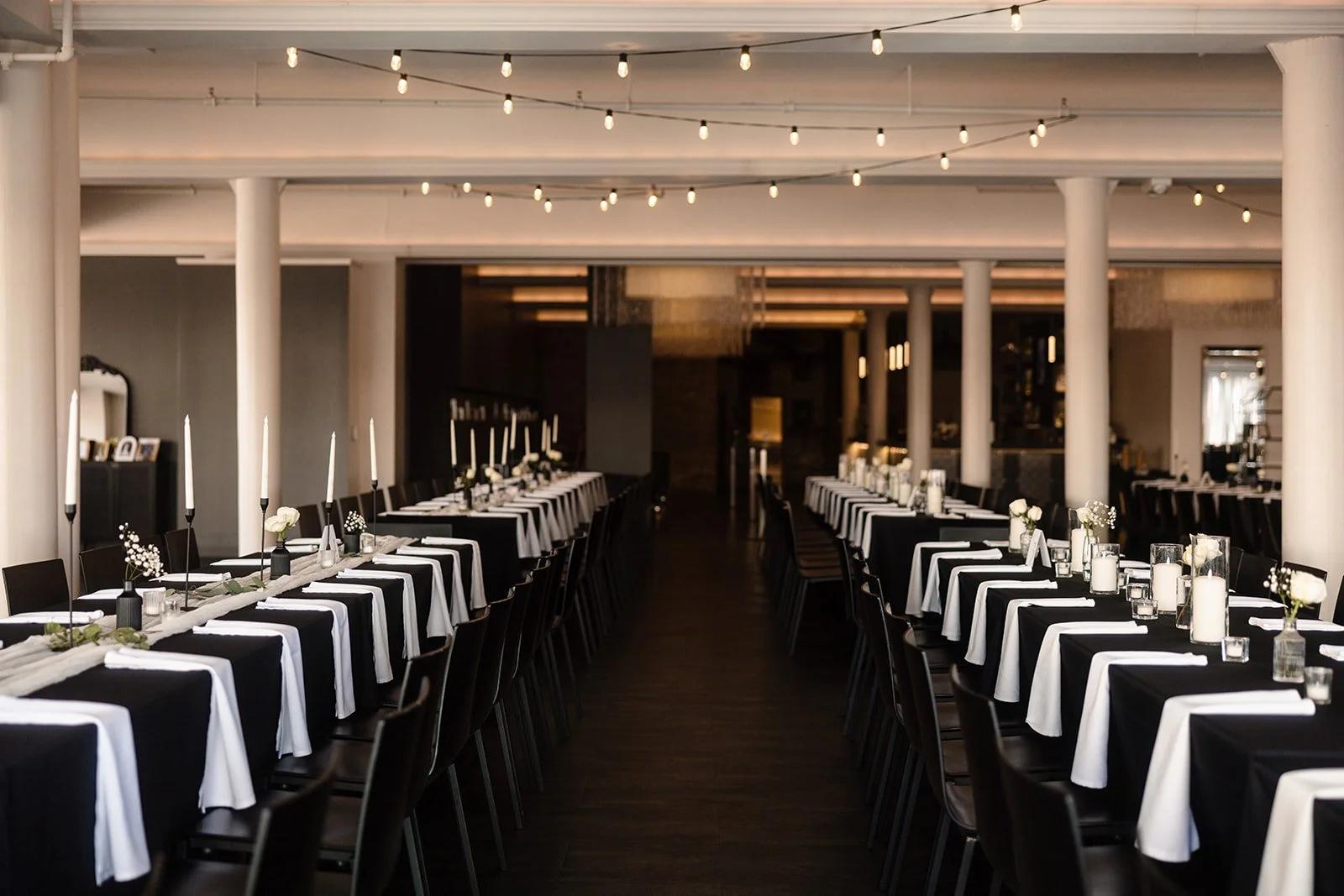 Elegant banquet room decorated with black and white tablecloths, tall white candles, small vases with white flowers and candles, black chairs, and string lights hanging from the ceiling.
