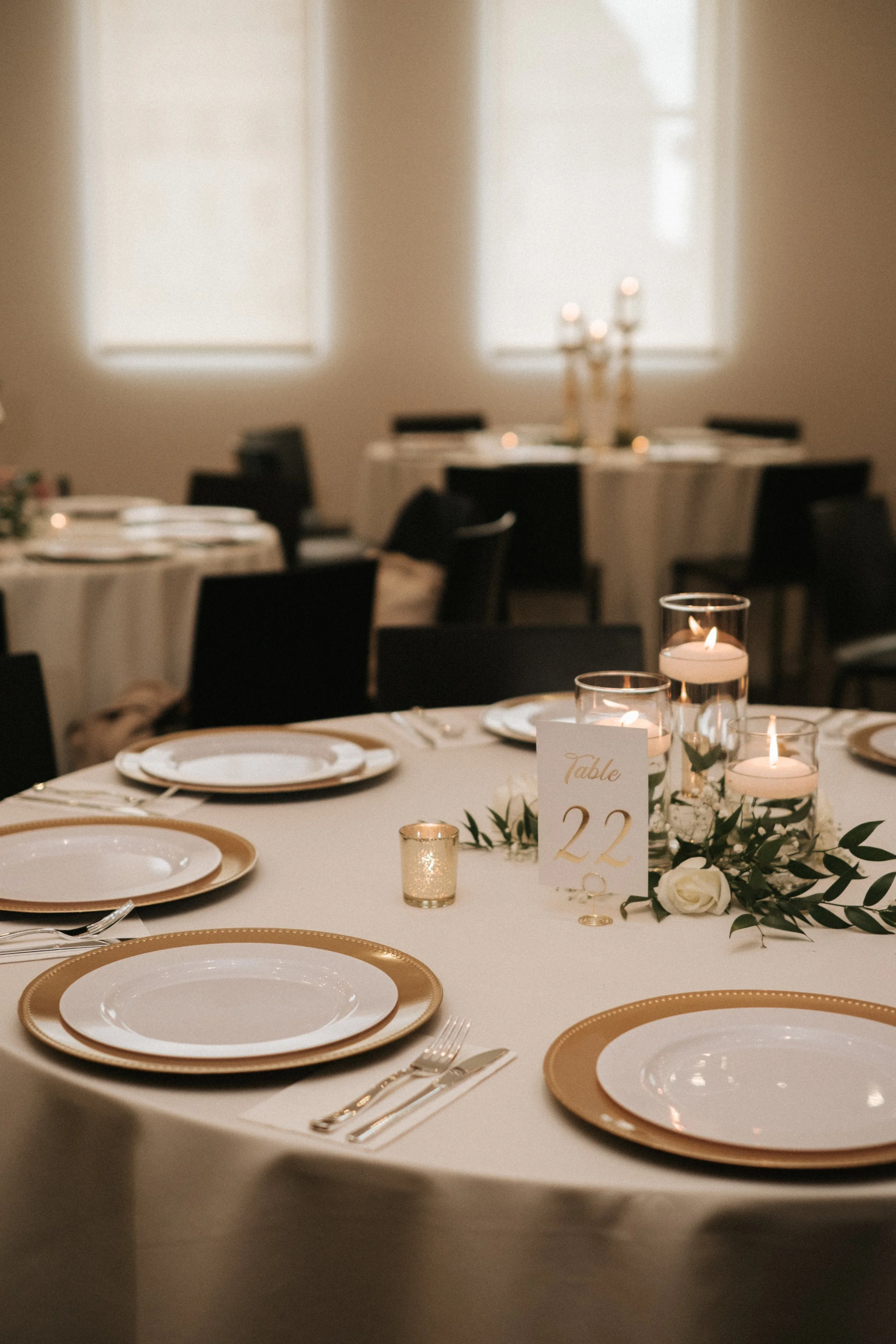 Elegant table setting with white and gold plates, silverware, glassware, candles, and a table number card, in a softly lit banquet room with windows and chairs.