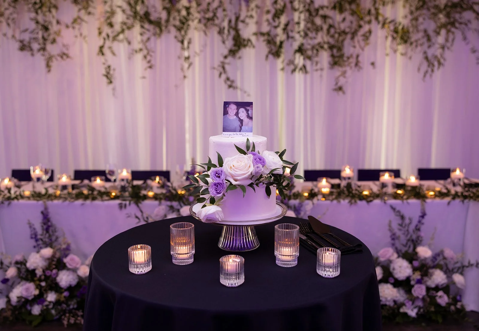 A wedding reception table with a small white cake decorated with flowers and a photo of a couple on top, surrounded by lit candles and floral arrangements, with a purple-lit backdrop.